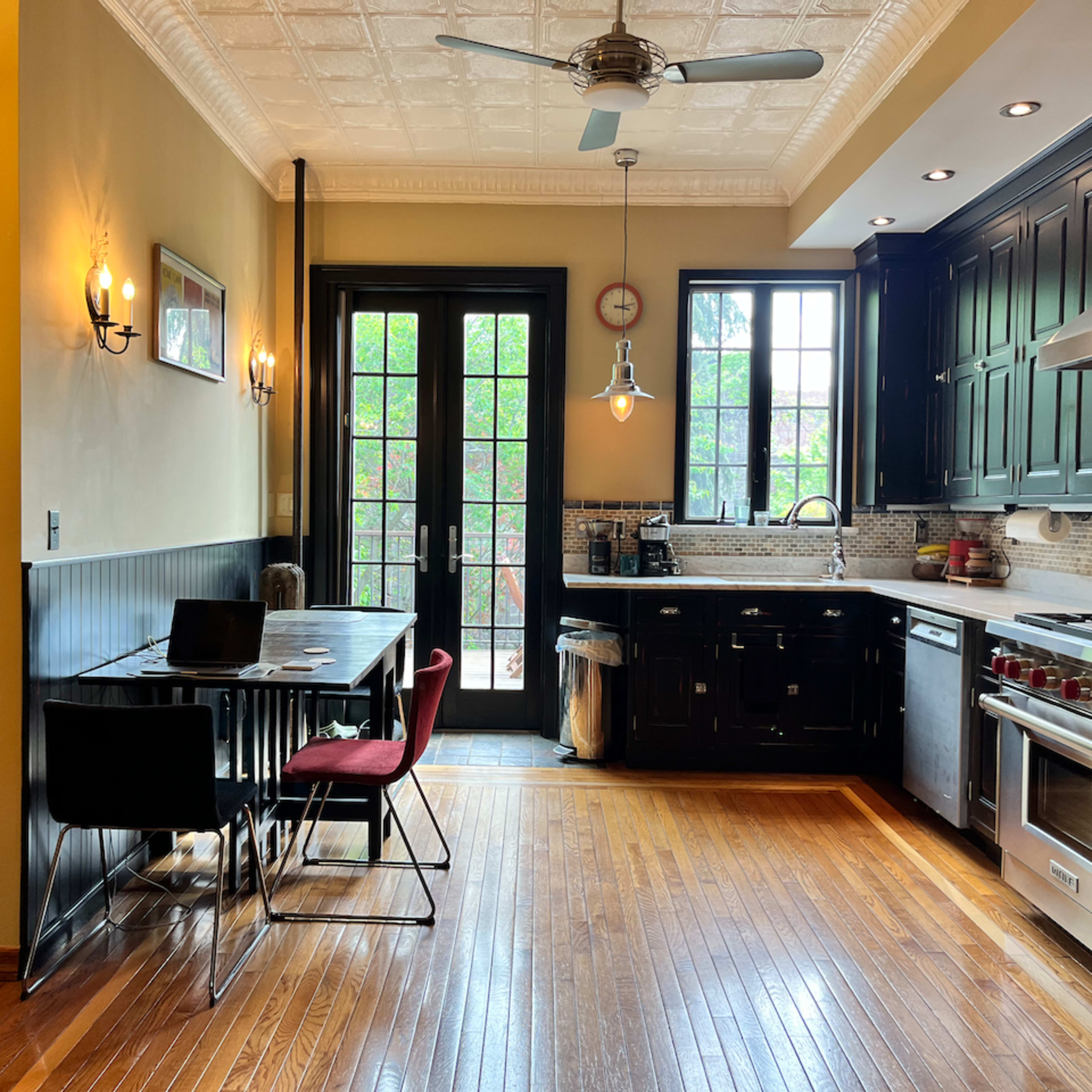 A kitchen with dark cabinets, stainless steel appliances, a dining table with black and red chairs, and large windows revealing a view of greenery outside.