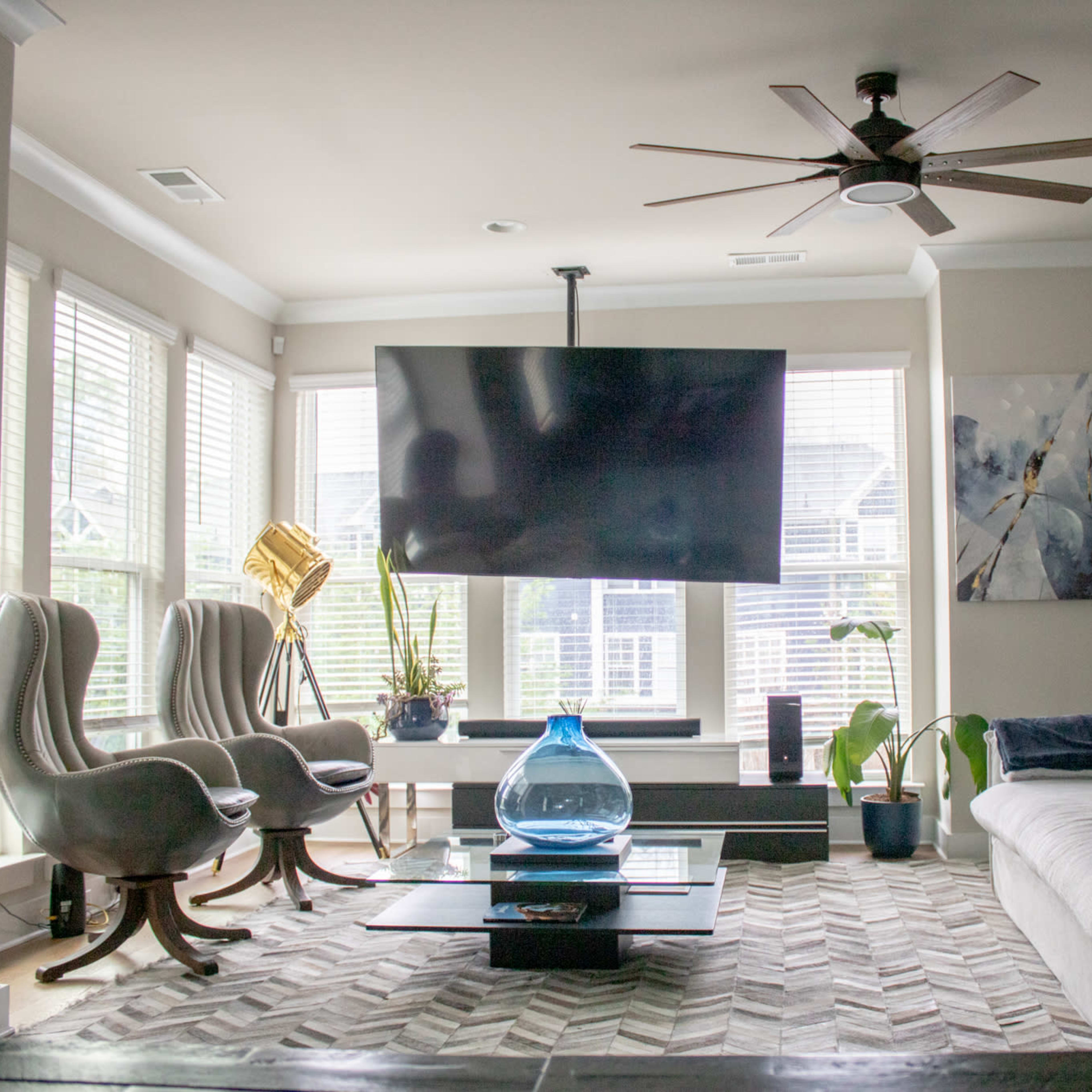 The living room features a large television mounted on the wall, two gray armchairs, a light-colored sectional sofa, and a coffee table with a blue glass vase, surrounded by large windows.