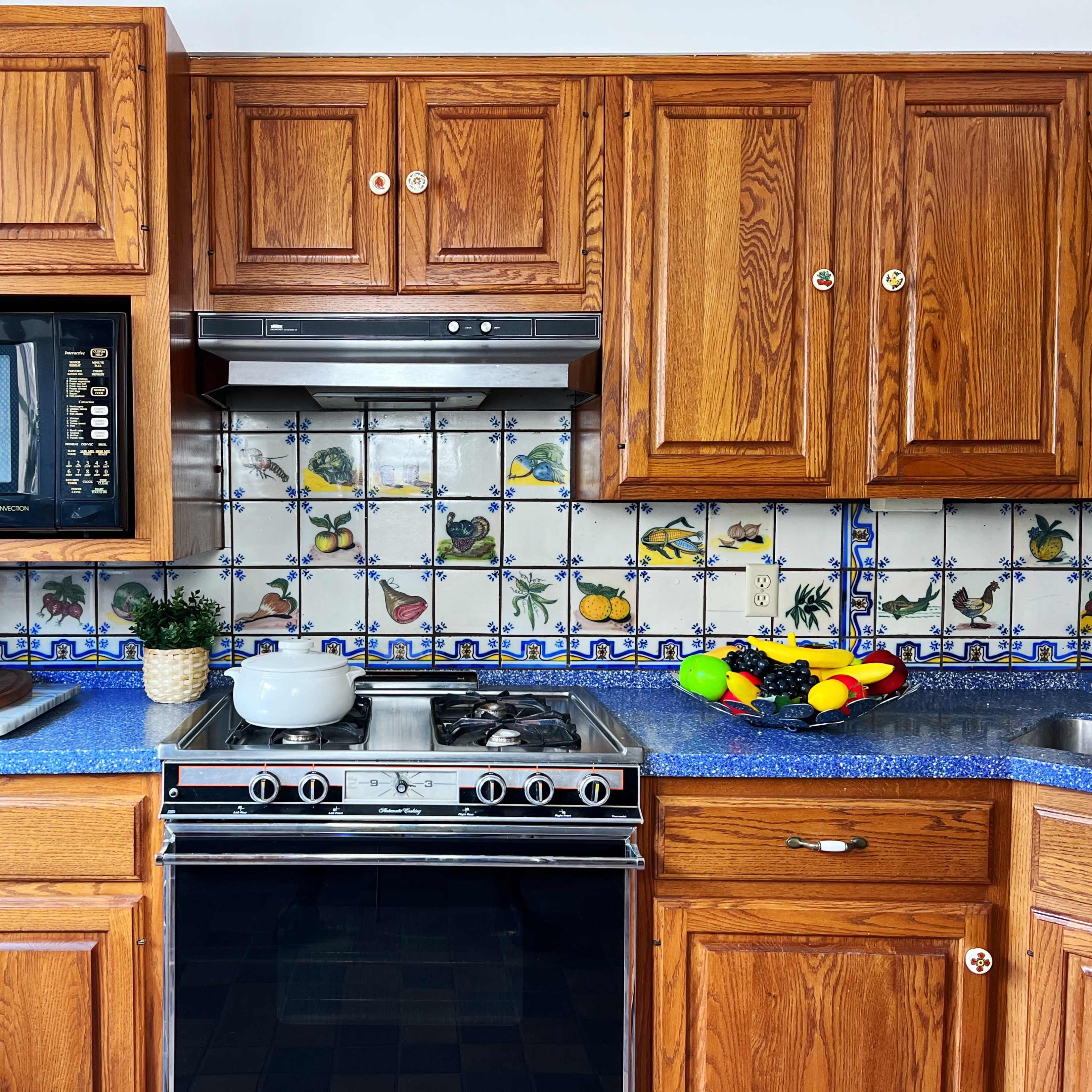 A wooden kitchen with a blue countertop, an oven, a microwave, and decorative tile backsplash featuring fruit designs.