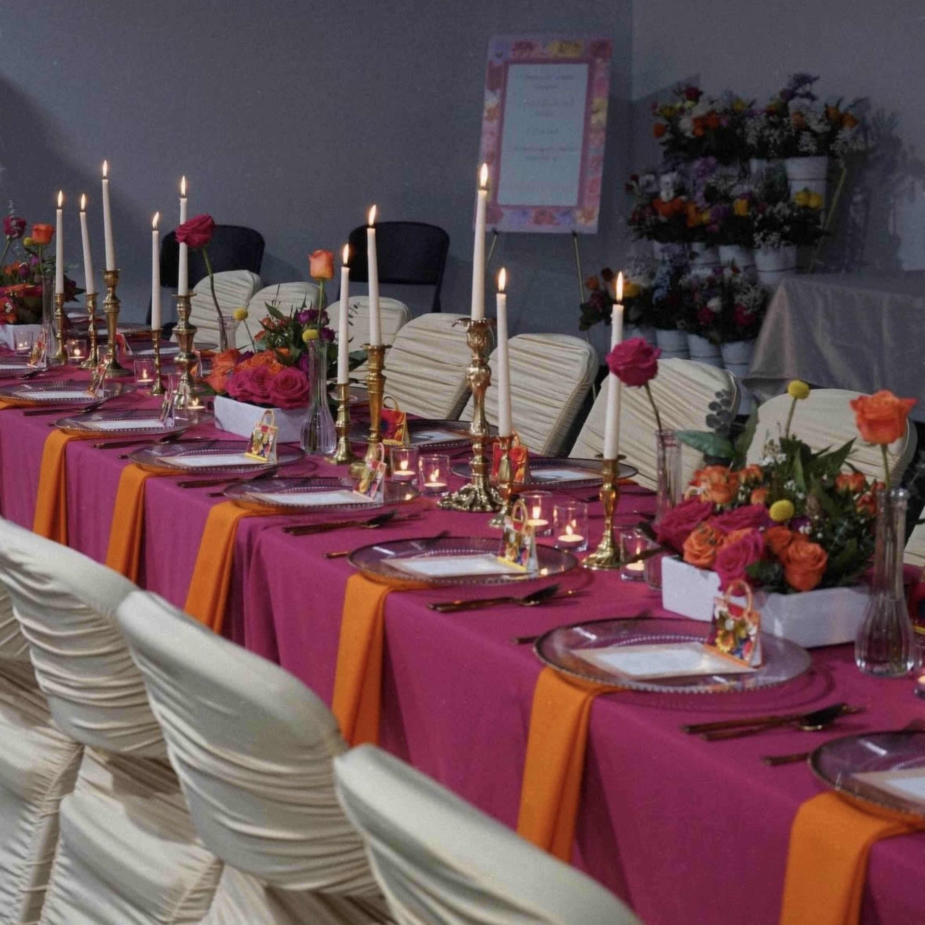 A long banquet table is set with pink and orange tablecloths, adorned with candles, floral centerpieces, and elegant place settings.