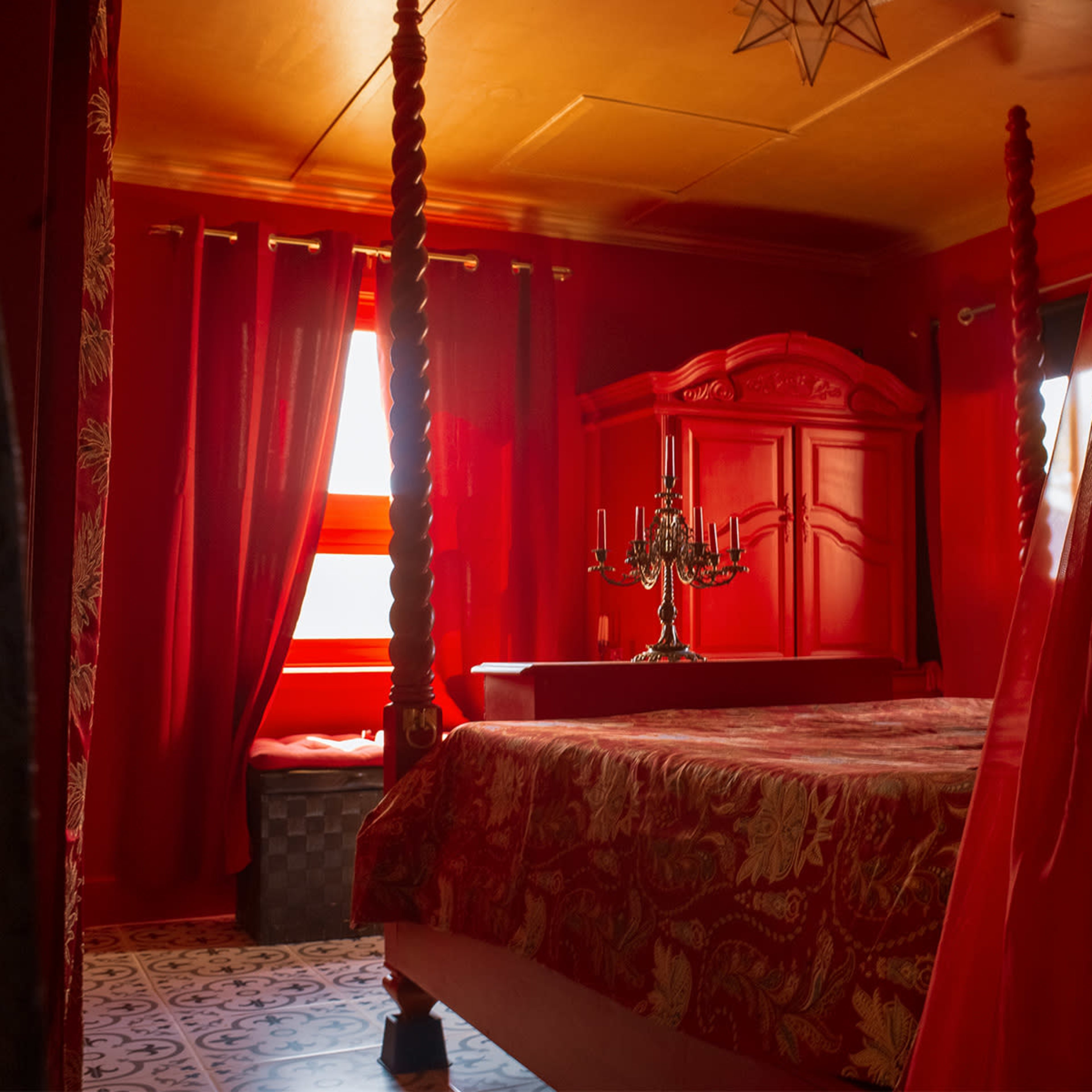A richly decorated bedroom with red walls and drapes, featuring a four-poster bed and a candelabra on a table.
