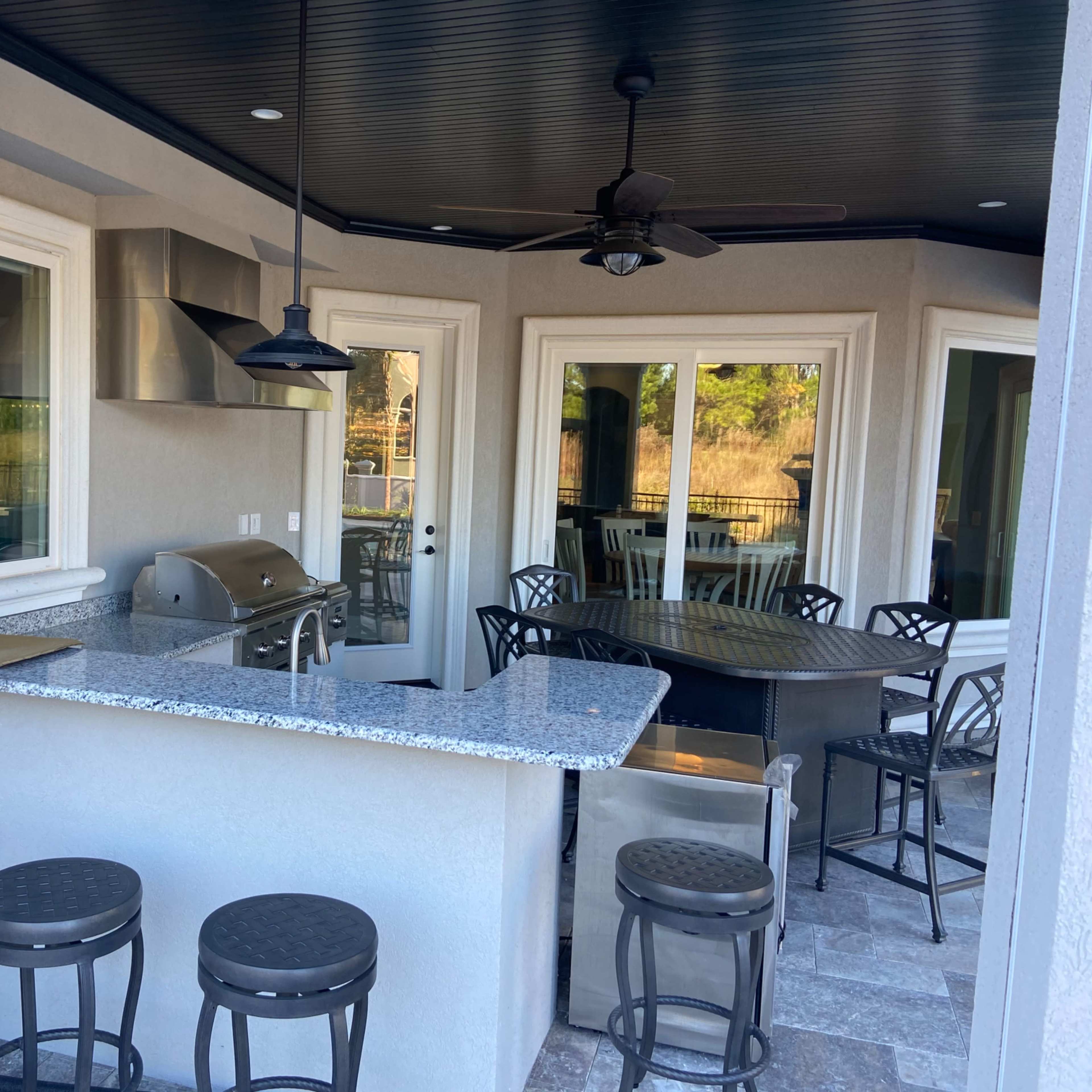 A covered outdoor kitchen and dining area featuring a granite bar, a grill, and a table with chairs.