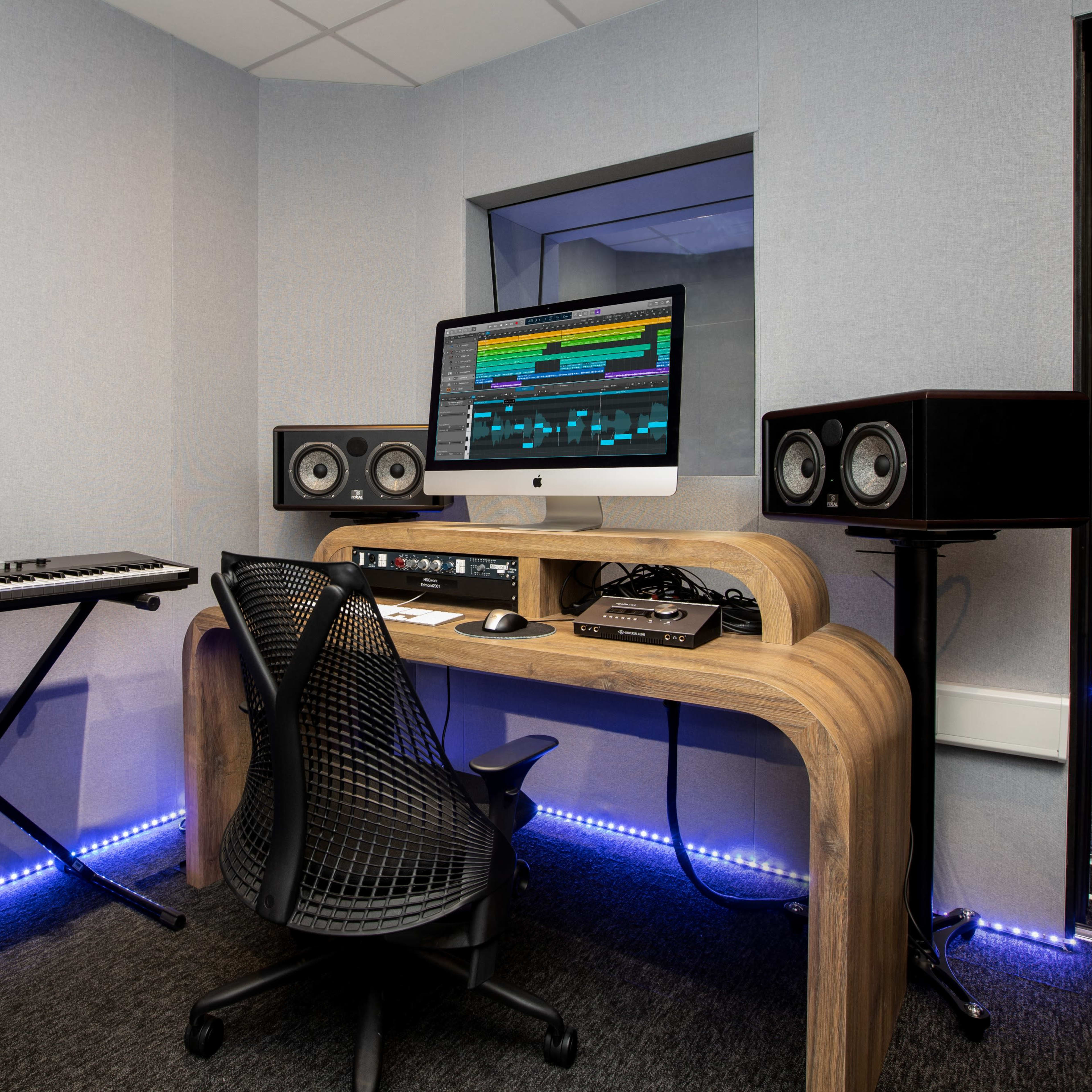 A music production studio featuring a computer desk with a monitor, two speakers, a keyboard, and a black ergonomic chair.