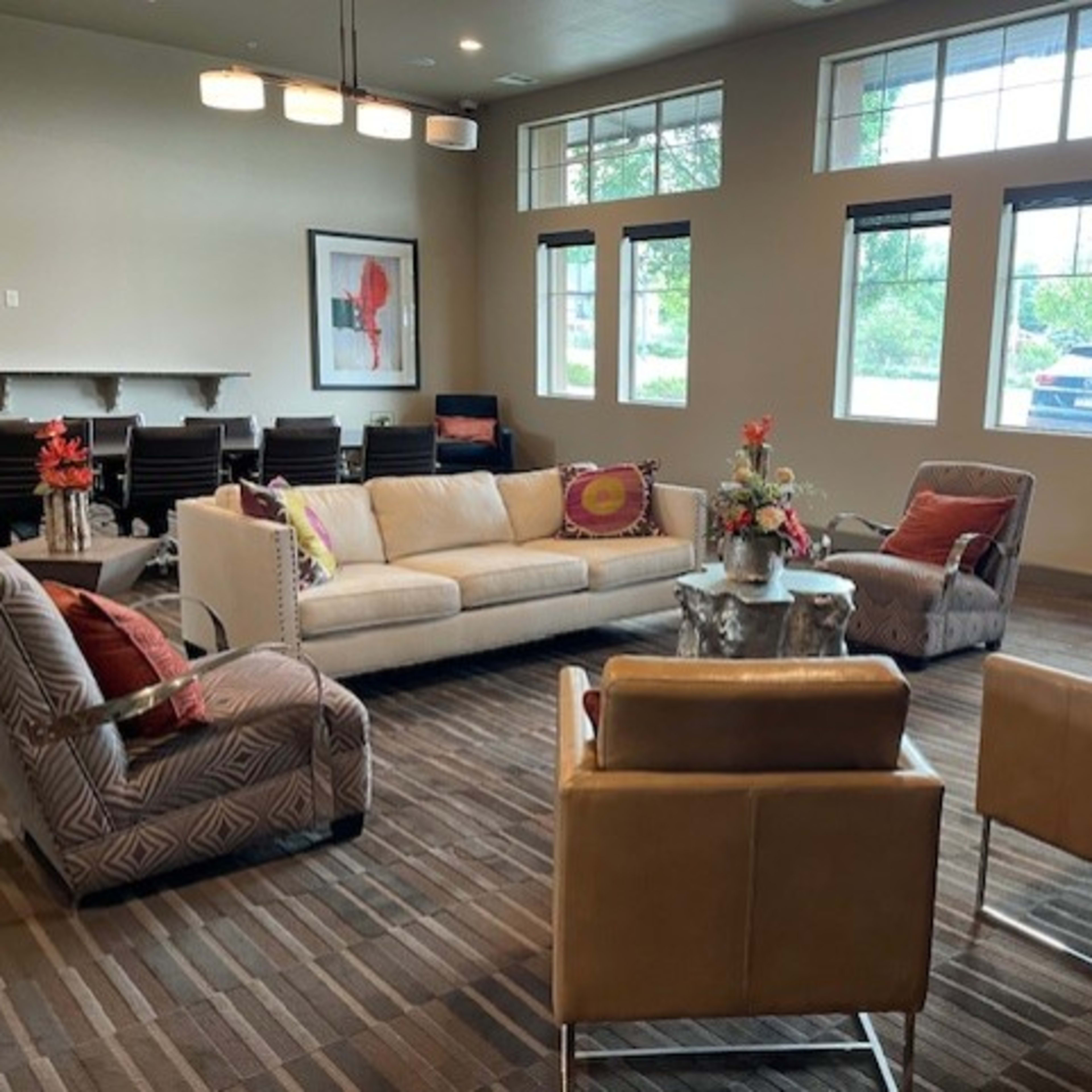 A modern lounge area with a white sofa, four armchairs, a small table with a floral arrangement, and large windows allowing natural light.