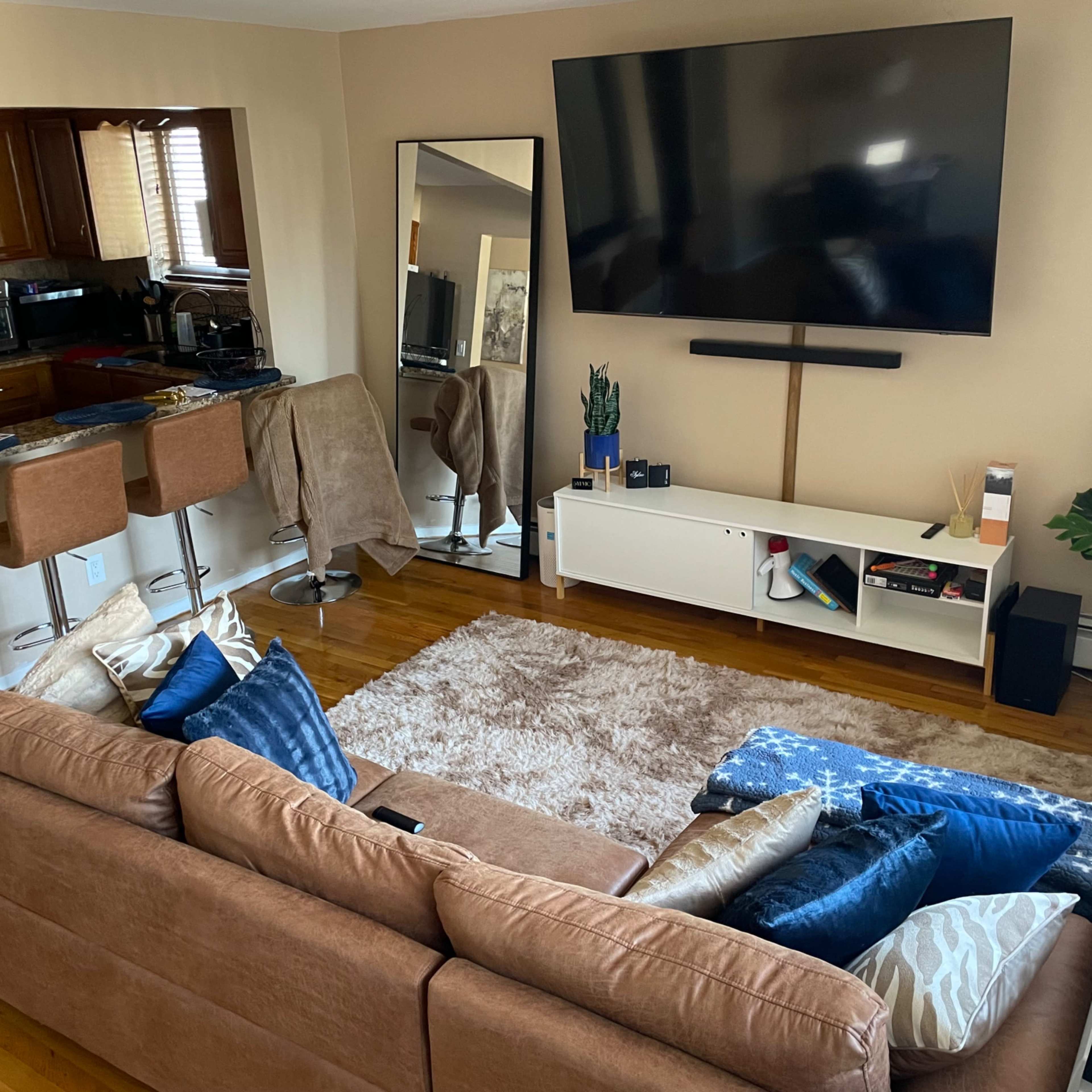 A living room features a brown sectional sofa with various blue cushions, a large wall-mounted TV, a coffee table, and a kitchen area in the background.