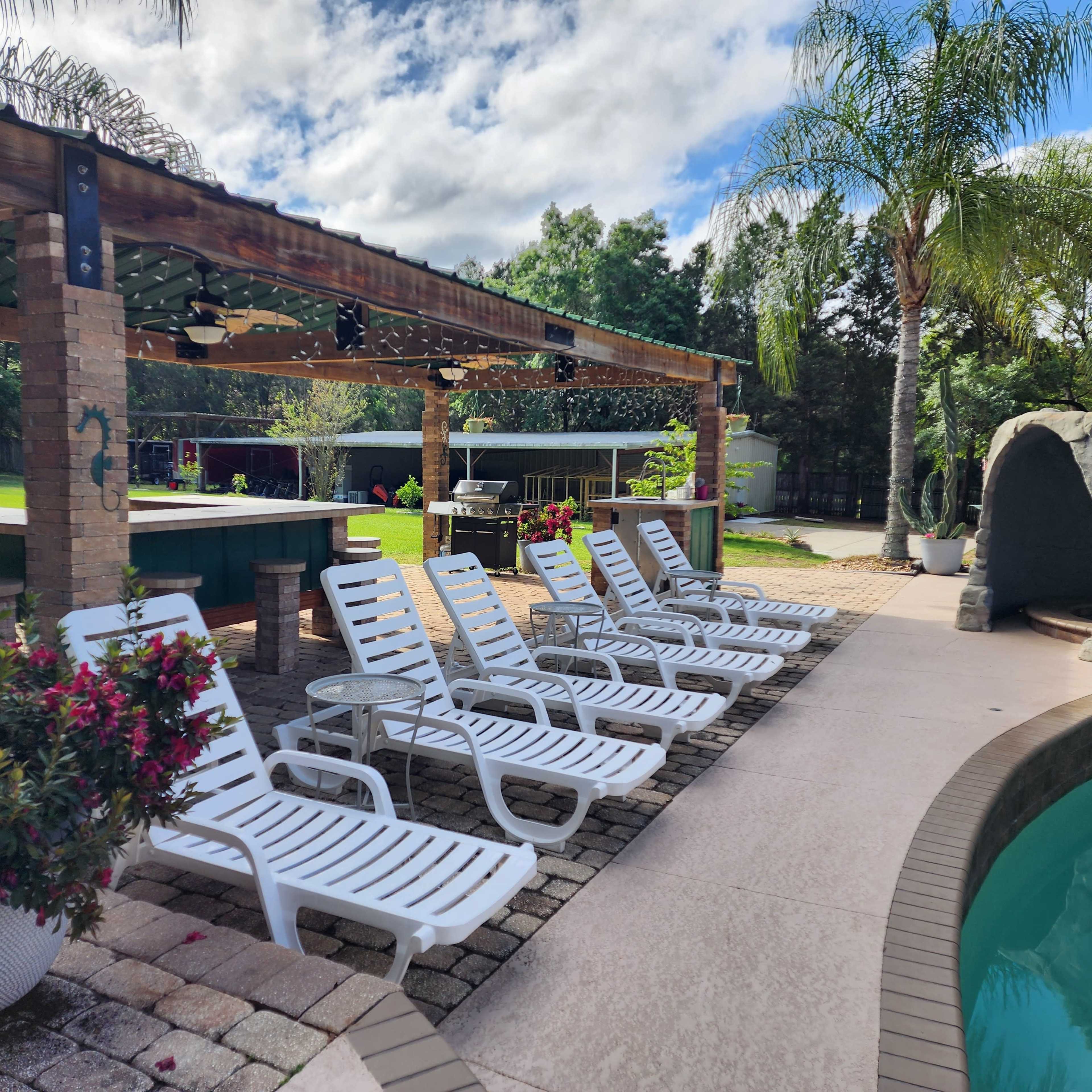 A row of white lounge chairs is arranged beside a swimming pool in a landscaped backyard with a gazebo and greenery.