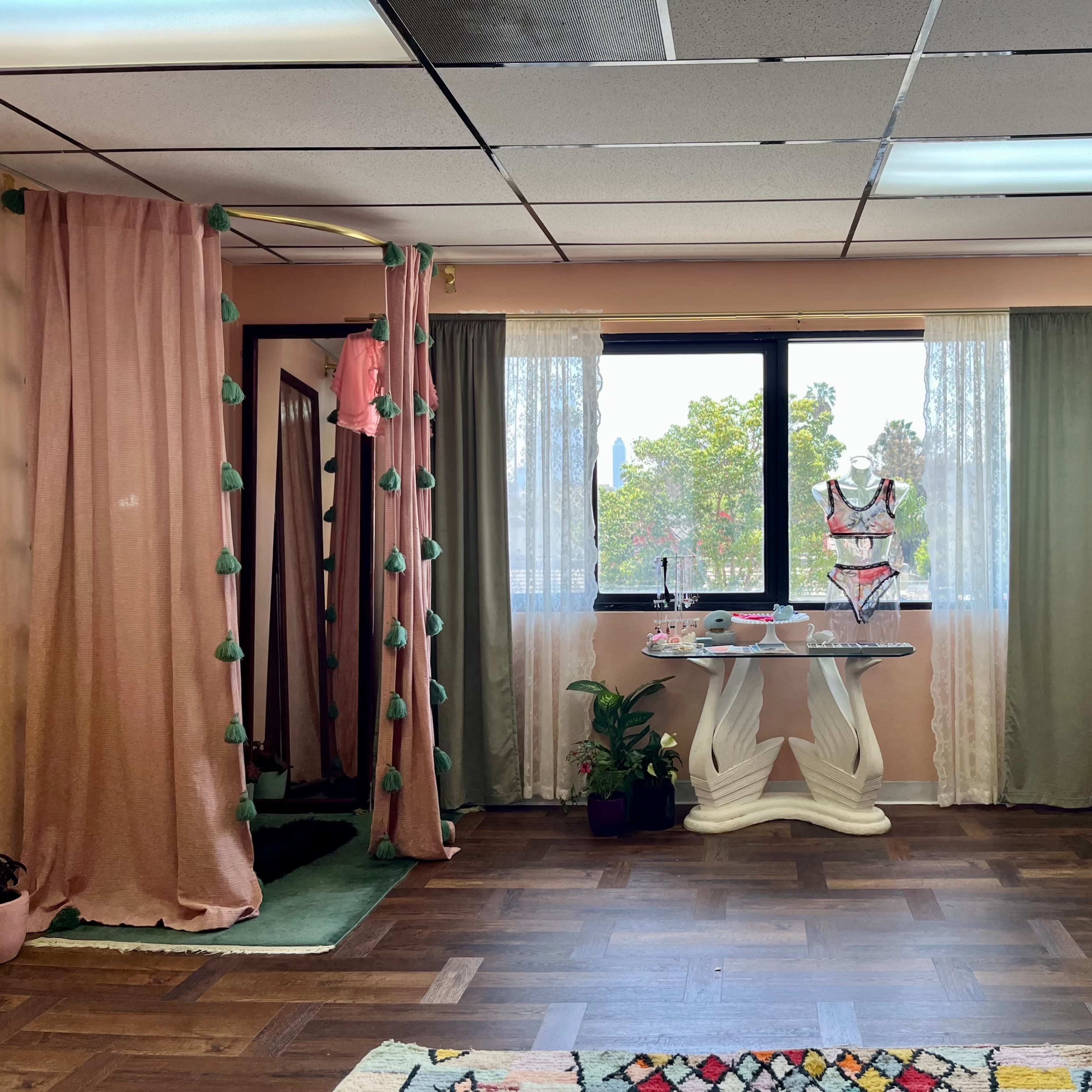 A boutique interior featuring a clothing rack with various garments, a fitting area with curtains, and a display table with lingerie near large windows.