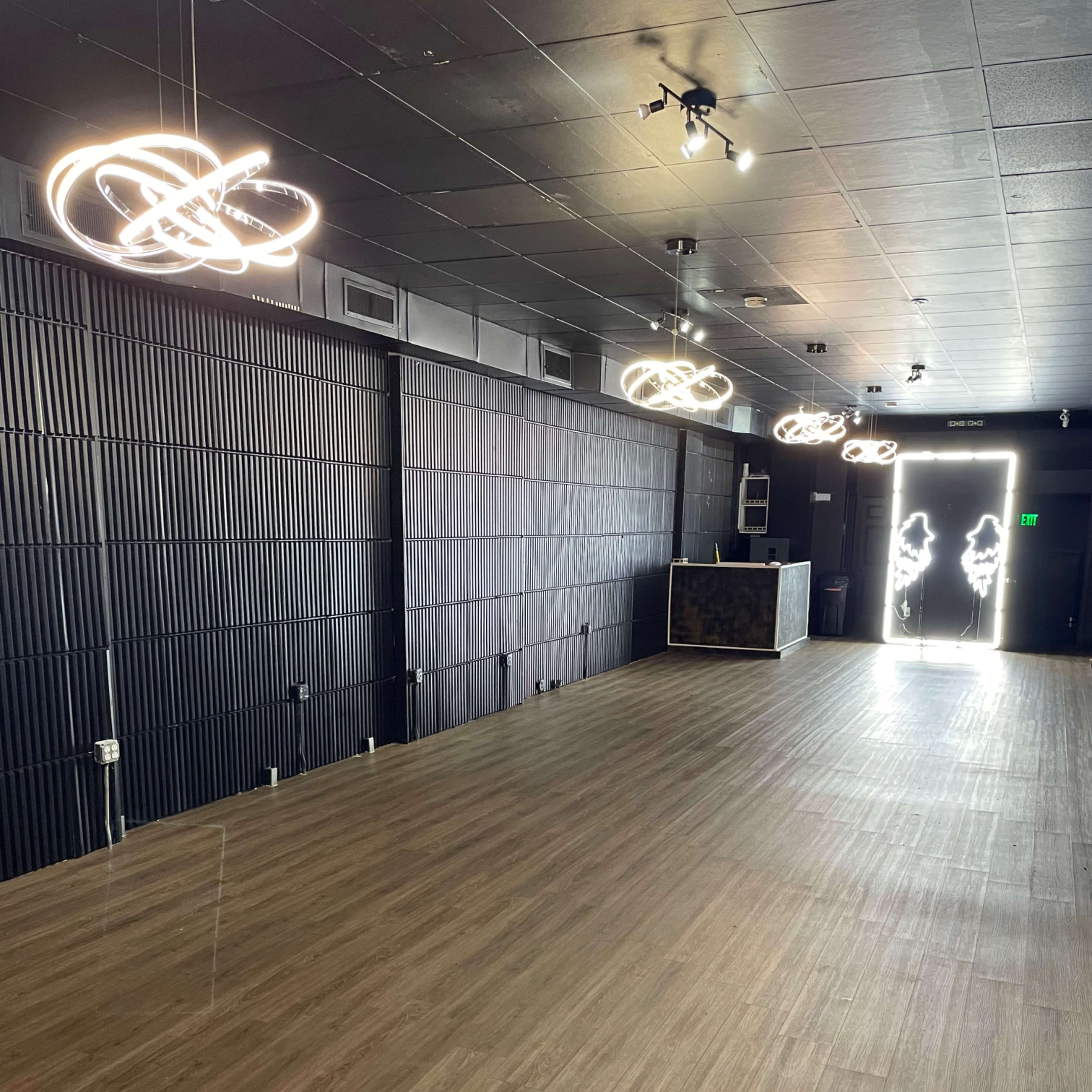 An empty room with wooden flooring, black walls, modern light fixtures, and an entrance to an outside space illuminated by bright light.