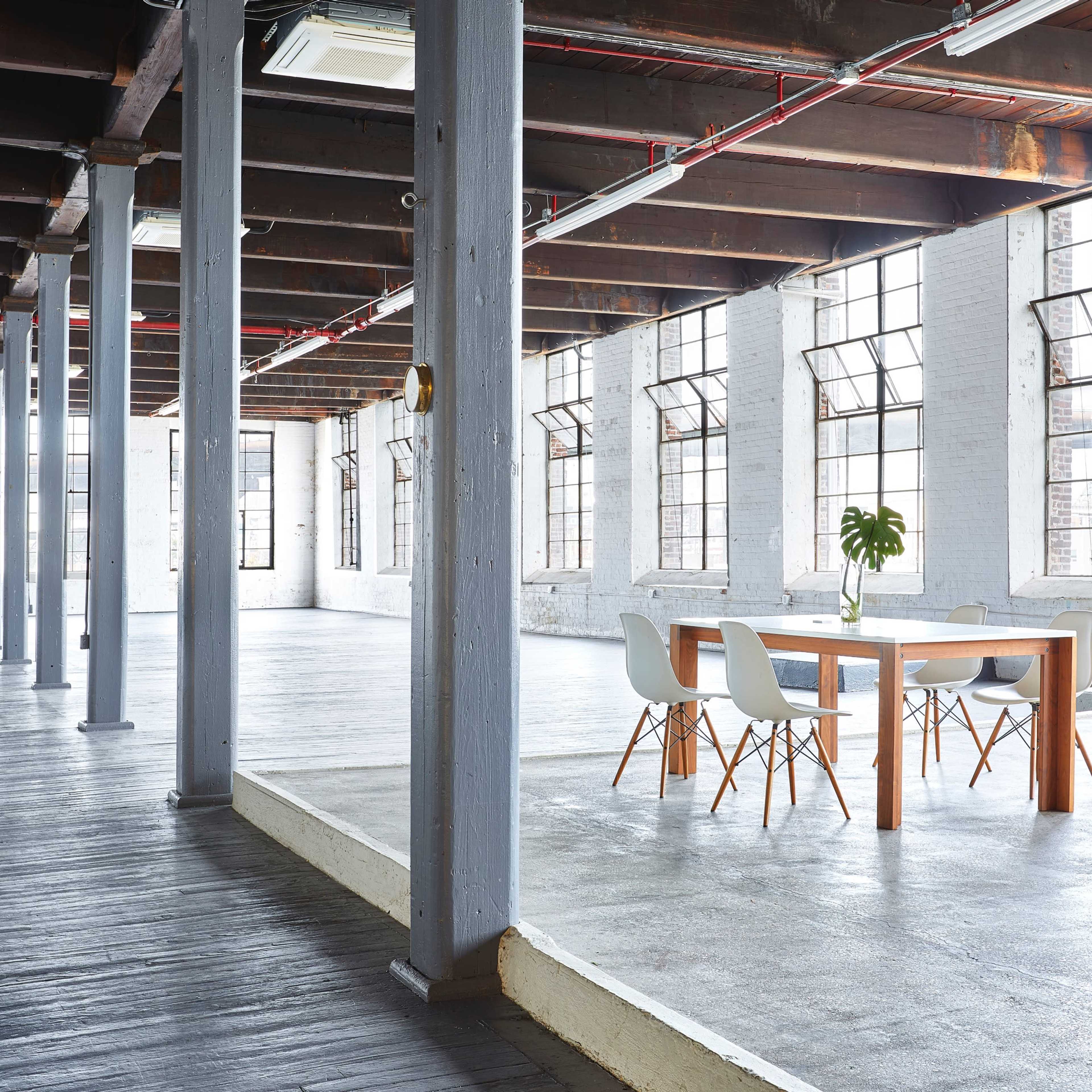 A spacious, industrial-style interior with large windows, wooden beams, and a dining table surrounded by chairs.