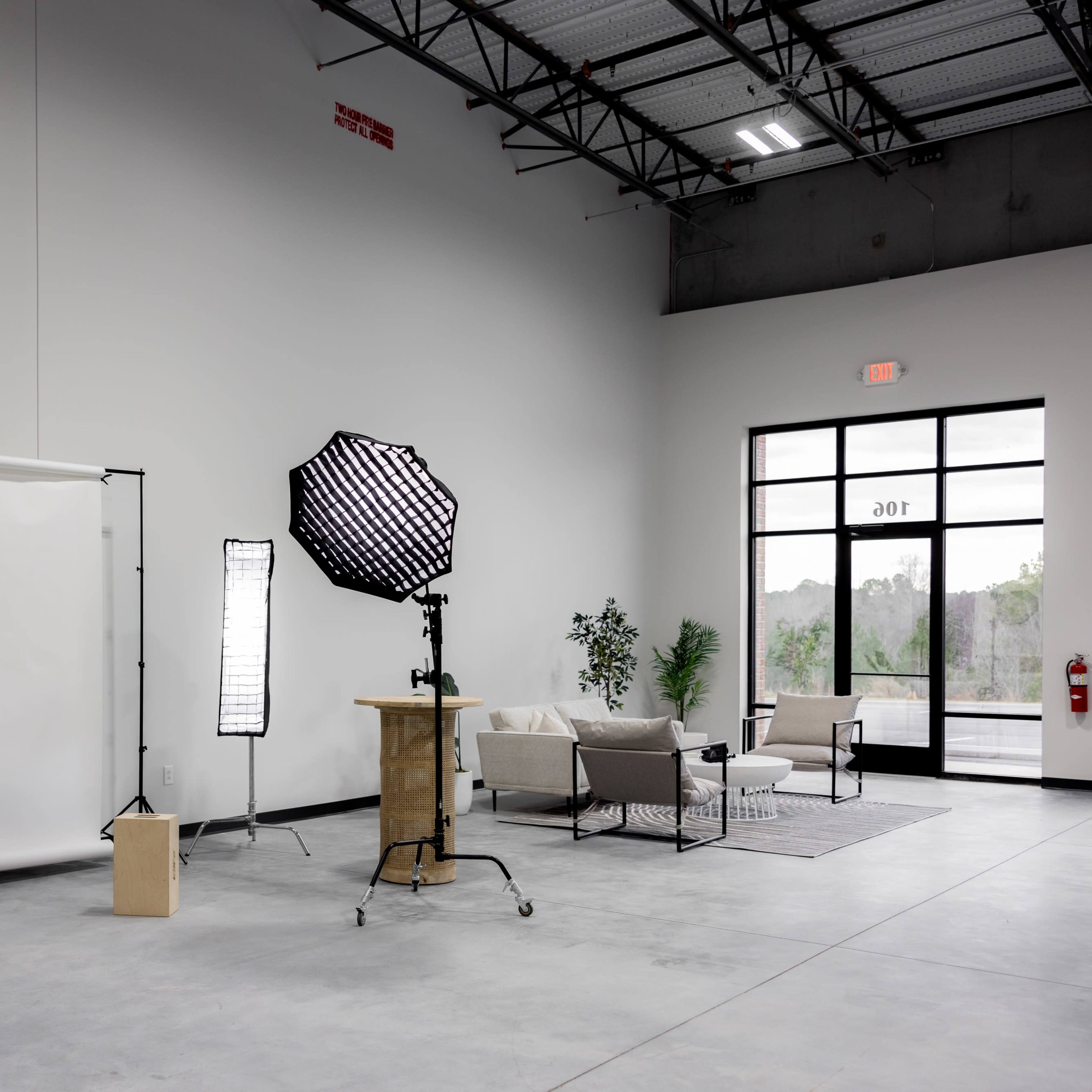 A spacious photography studio featuring a white backdrop, various lighting equipment, and a seating area.