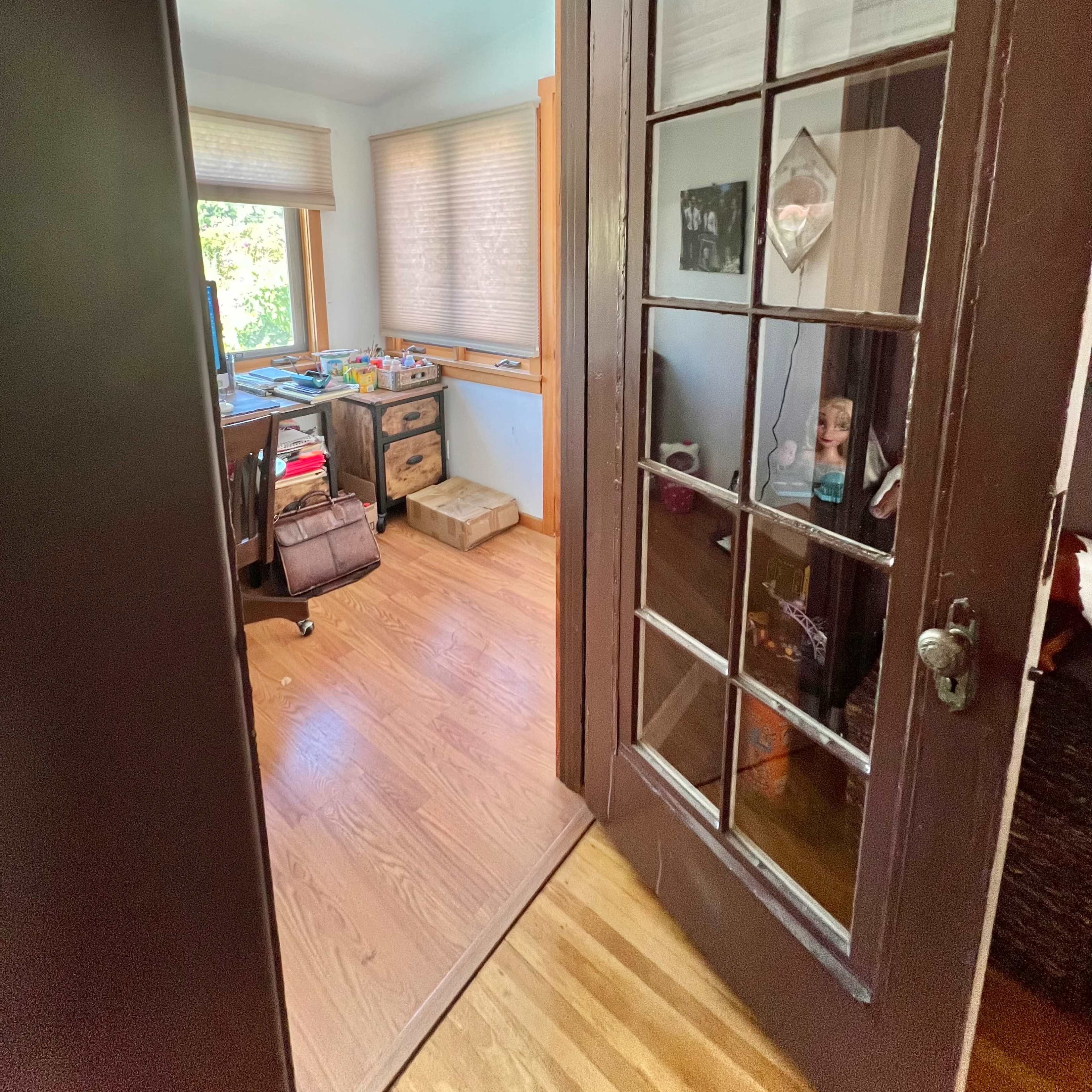 An open door leading into a brightly lit room with wooden floors, a desk cluttered with items, and a window with sheer shades.