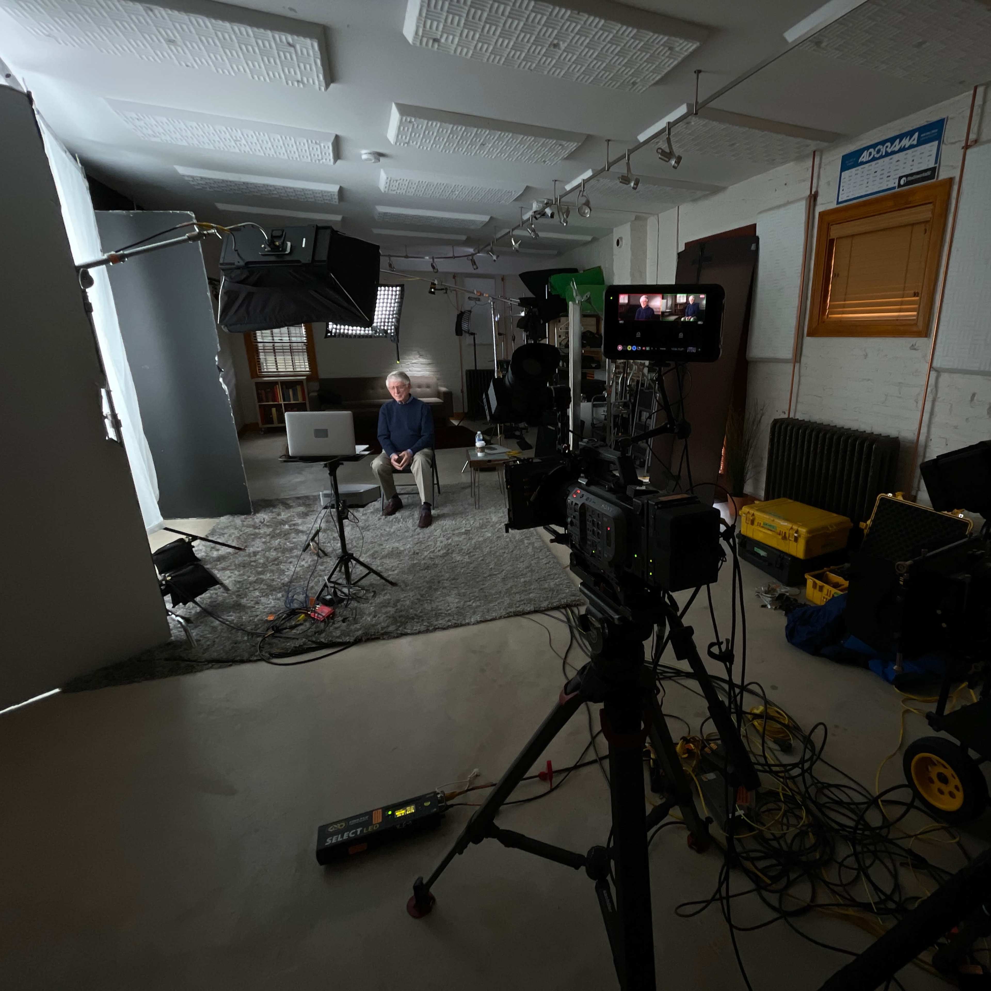 A man sits in front of a camera setup in a studio with various lighting equipment and cables visible around him.