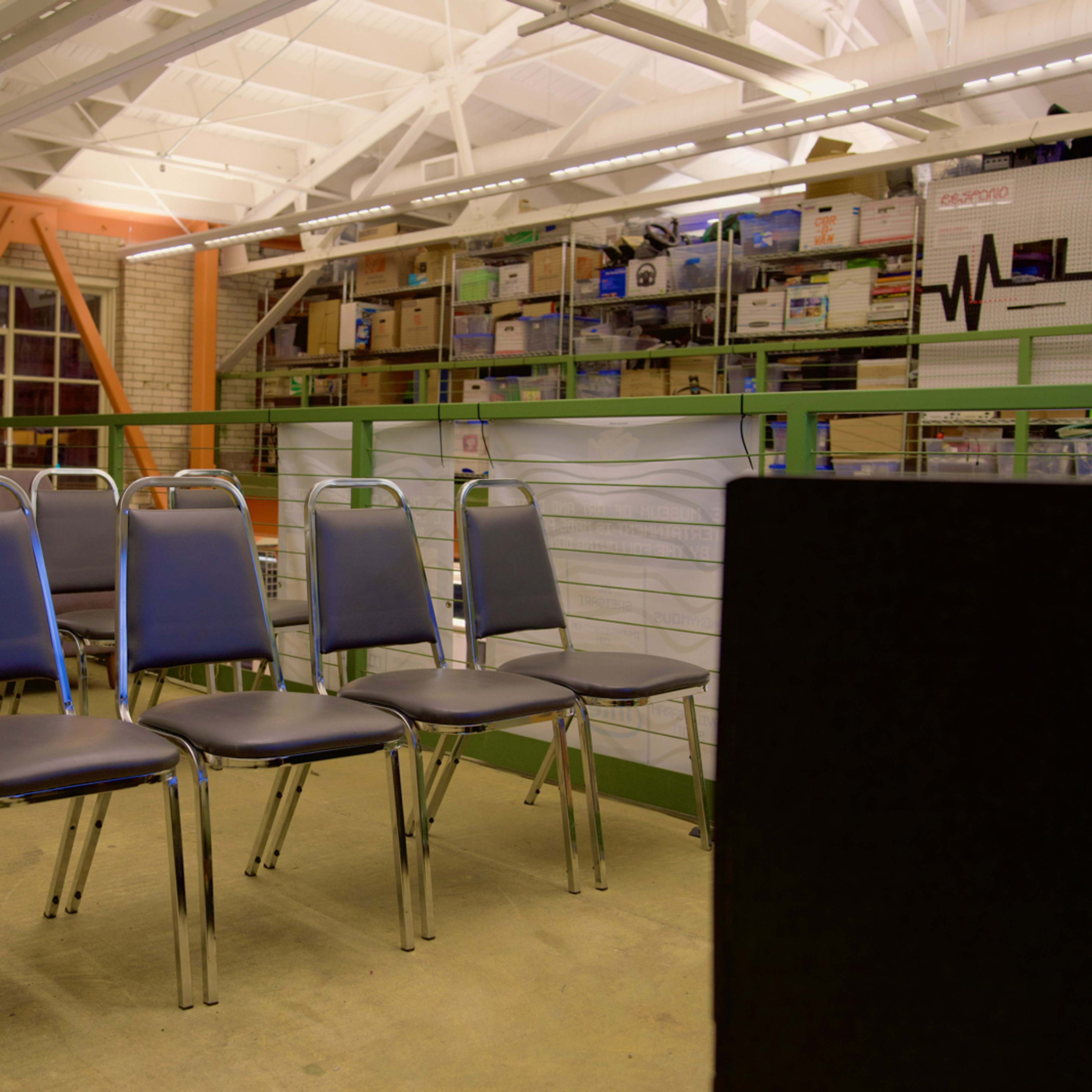 A row of metal chairs is lined up in a brightly lit, industrial-style room filled with shelves of boxes.