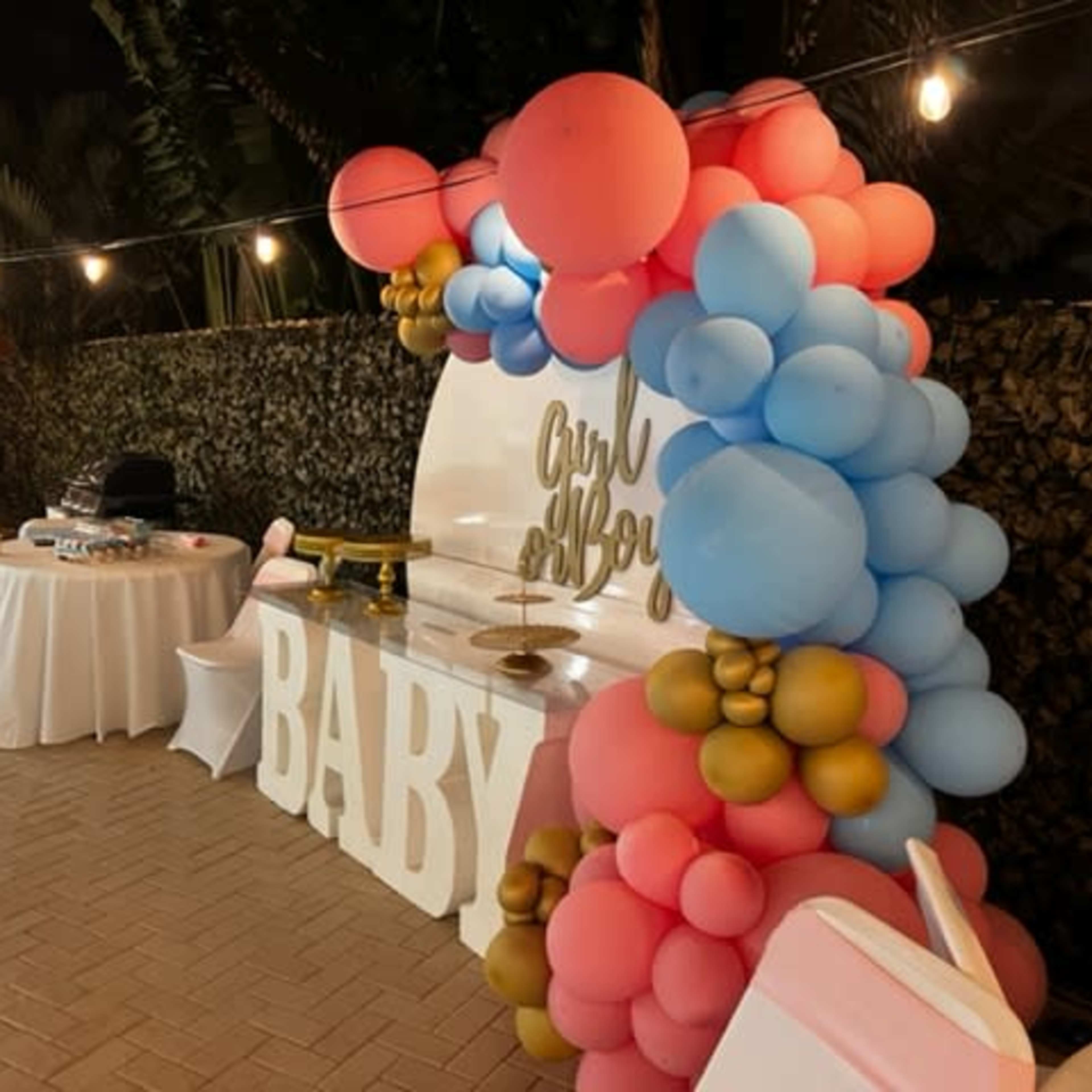 A decorated area near a pool, featuring a balloon arch in pink, blue, and gold, with a table set up for a celebration labeled "BABY."