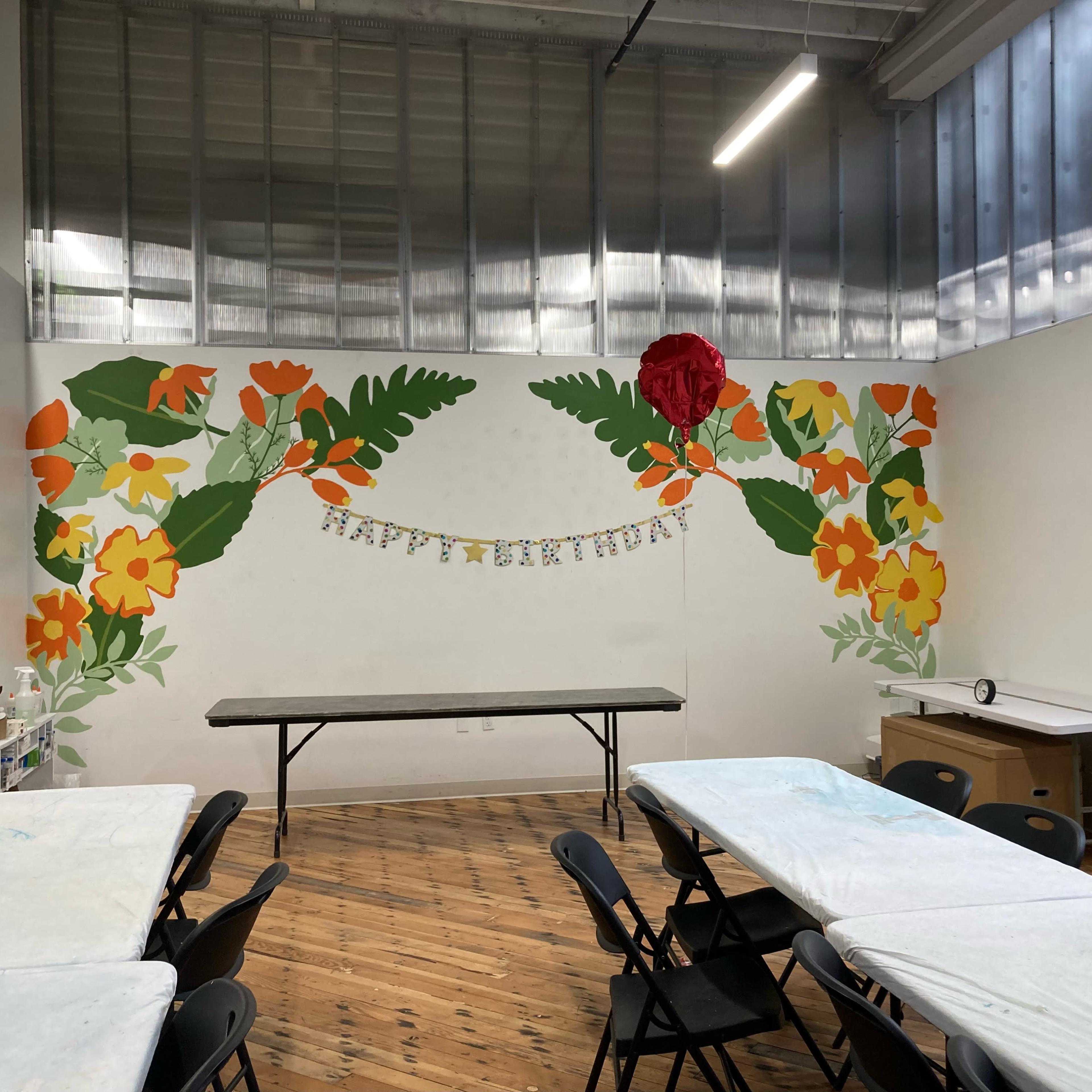 A brightly decorated room with a mural of flowers and leaves on one wall, a long table in the center, and folding chairs arranged around it.