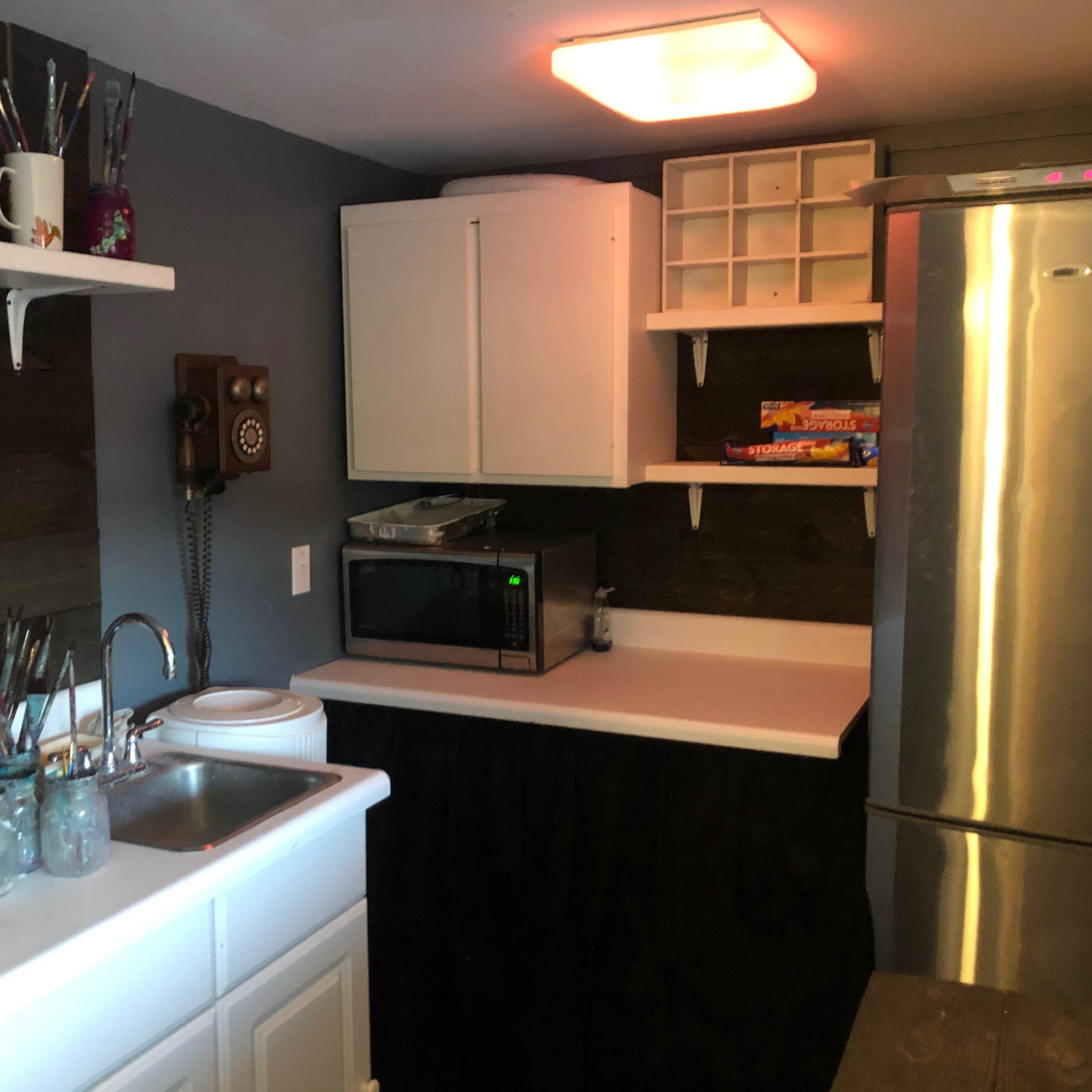 A small kitchen area with a sink, microwave, refrigerator, and shelves holding various items and art supplies.