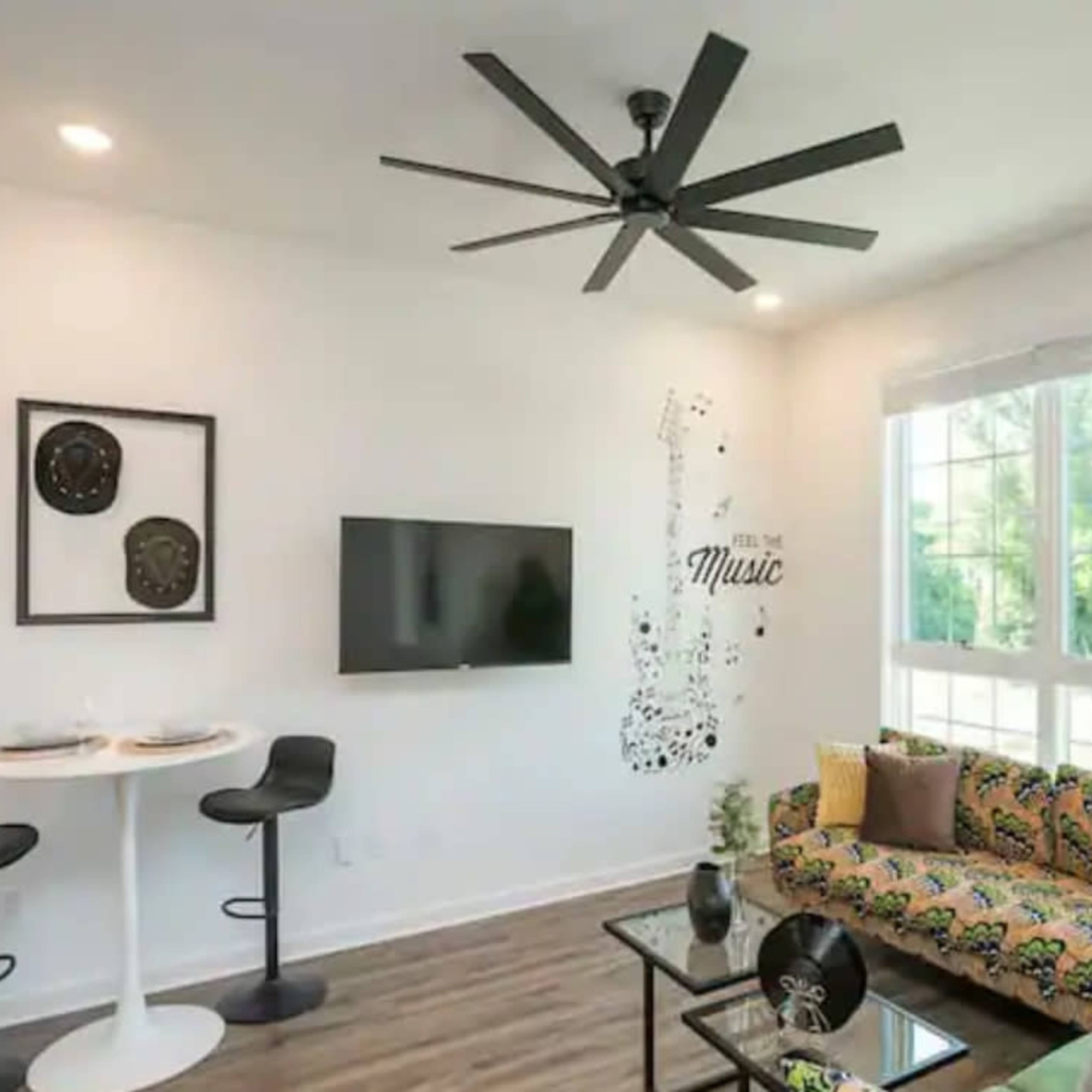 A modern living area with a green patterned sofa, a round white dining table with two stools, a wall-mounted TV, and a decorative wall design featuring musical notes.