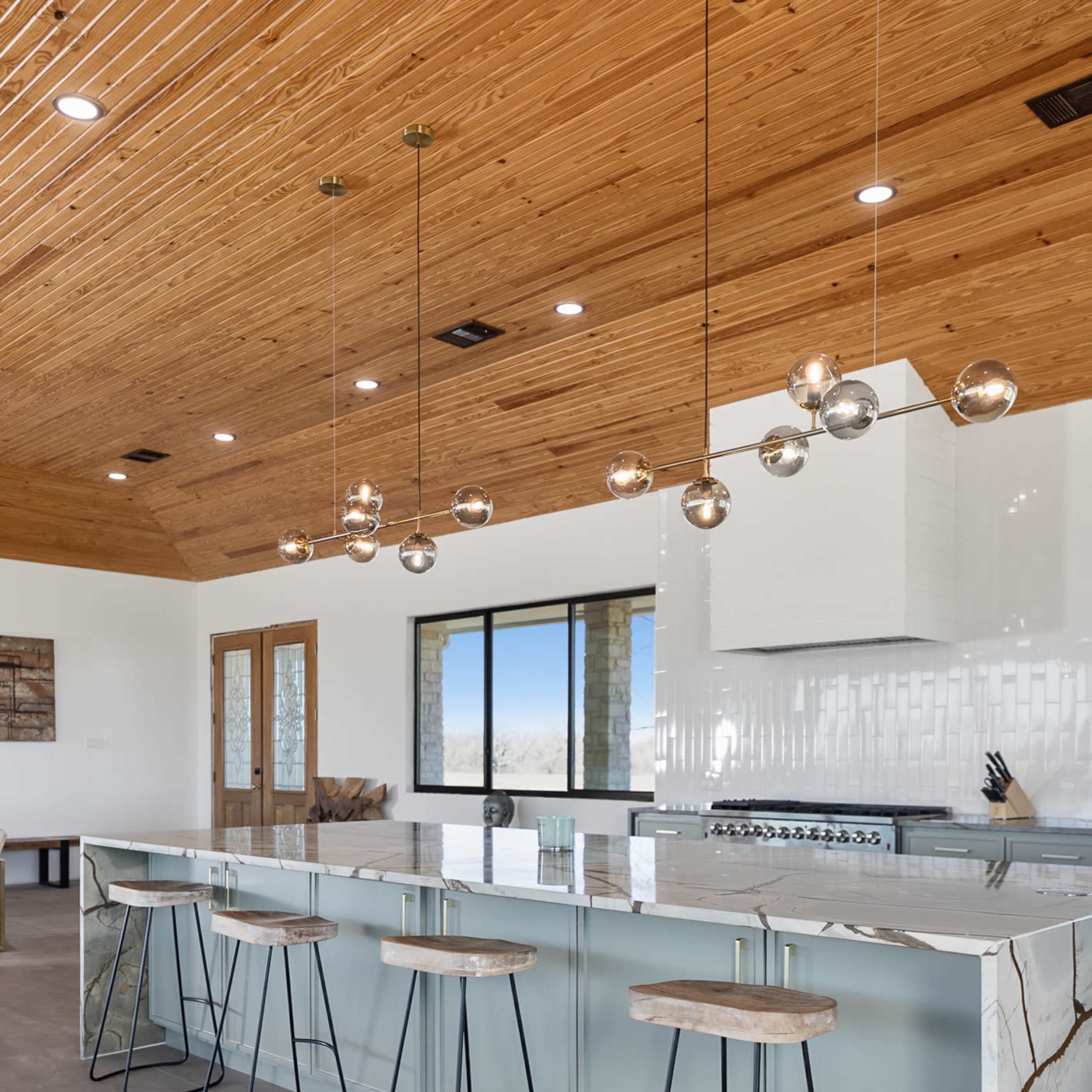 A modern kitchen with a large island featuring a marble countertop and four bar stools, under a wooden ceiling with pendant lights.