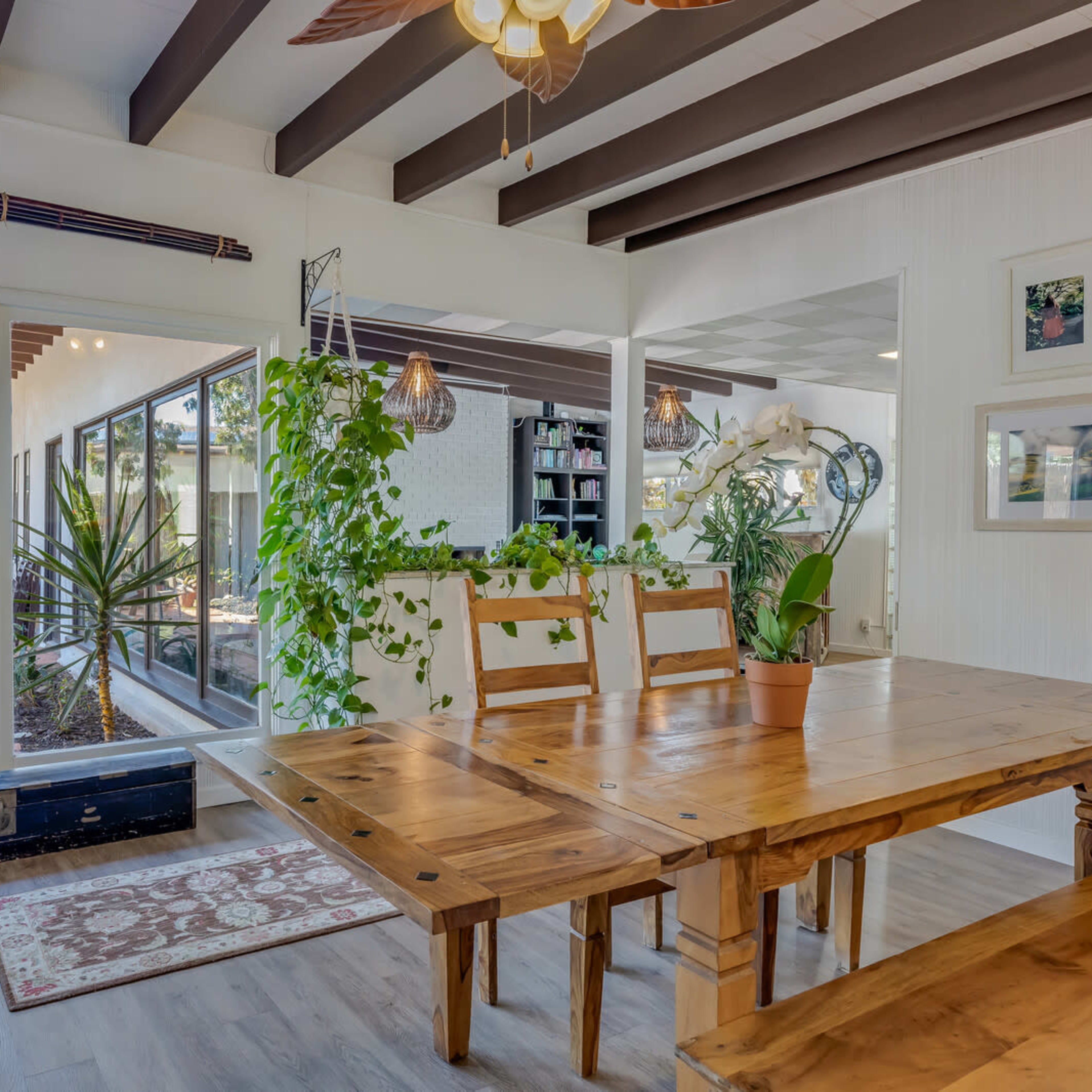 A wooden dining table with chairs is positioned near large glass doors that open to a patio filled with plants and outdoor furniture.