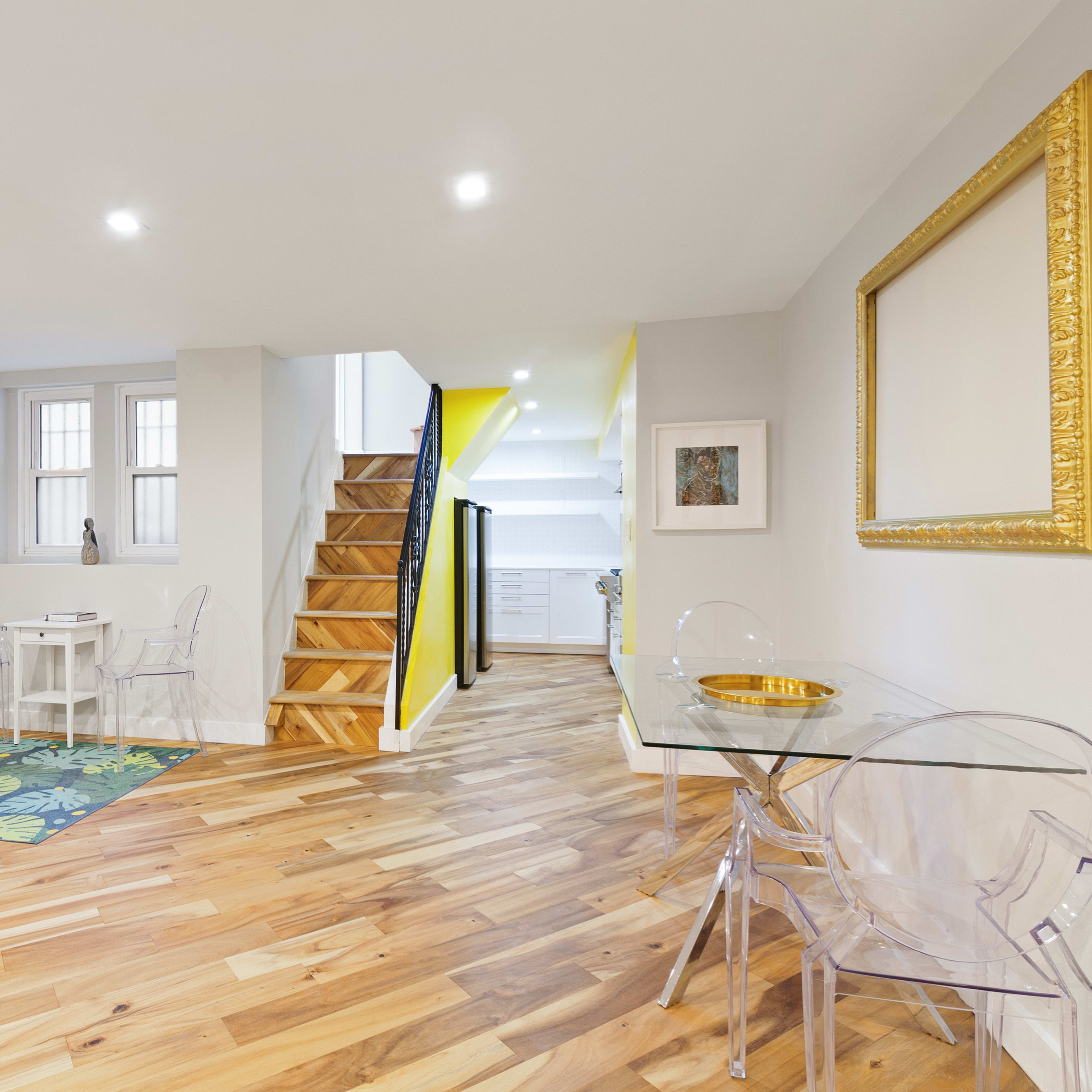 A bright, modern living space with wooden flooring, a staircase leading to an upper level, and a small dining area featuring transparent chairs.