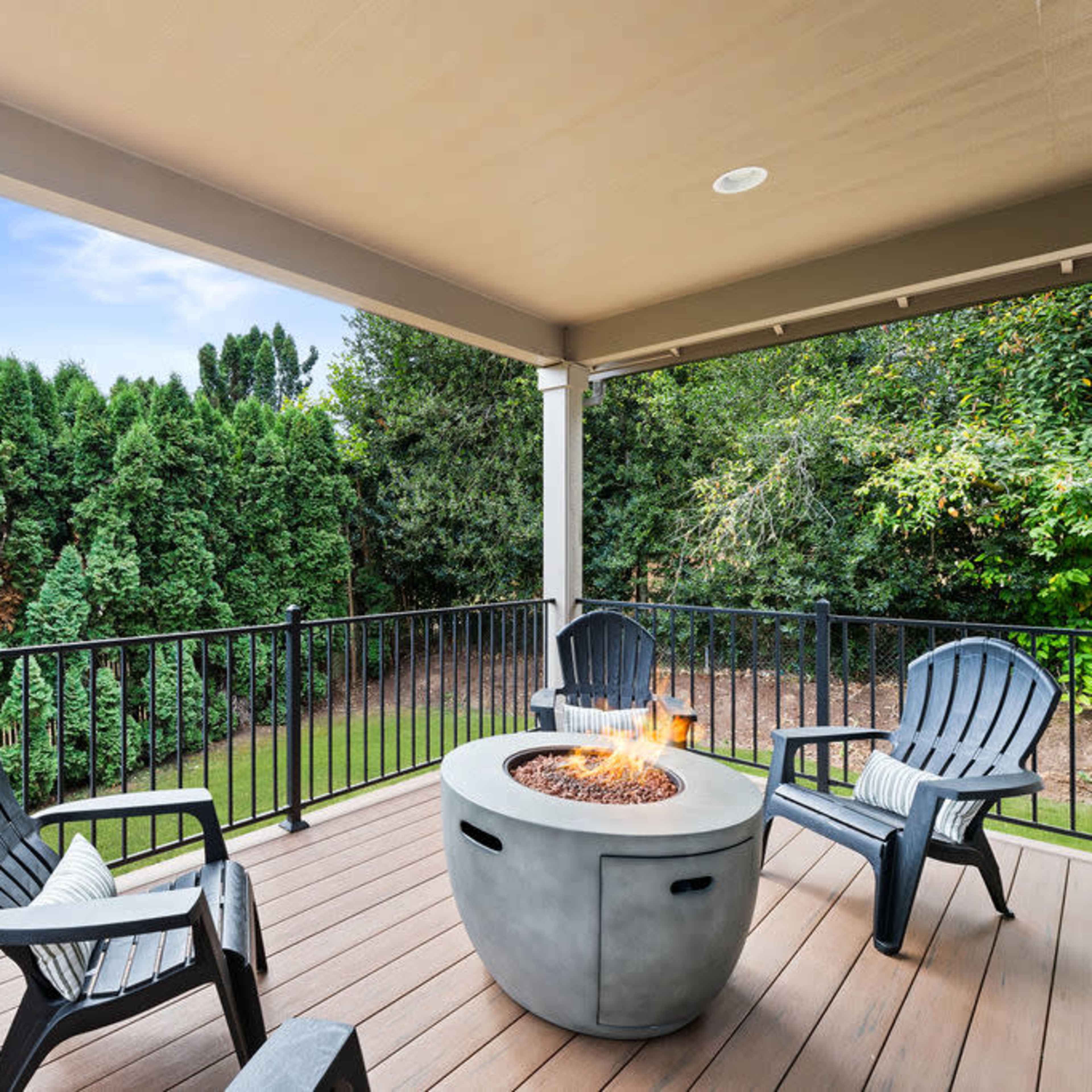 A covered balcony features three black Adirondack chairs arranged around a fire pit, with a green backyard visible in the background.