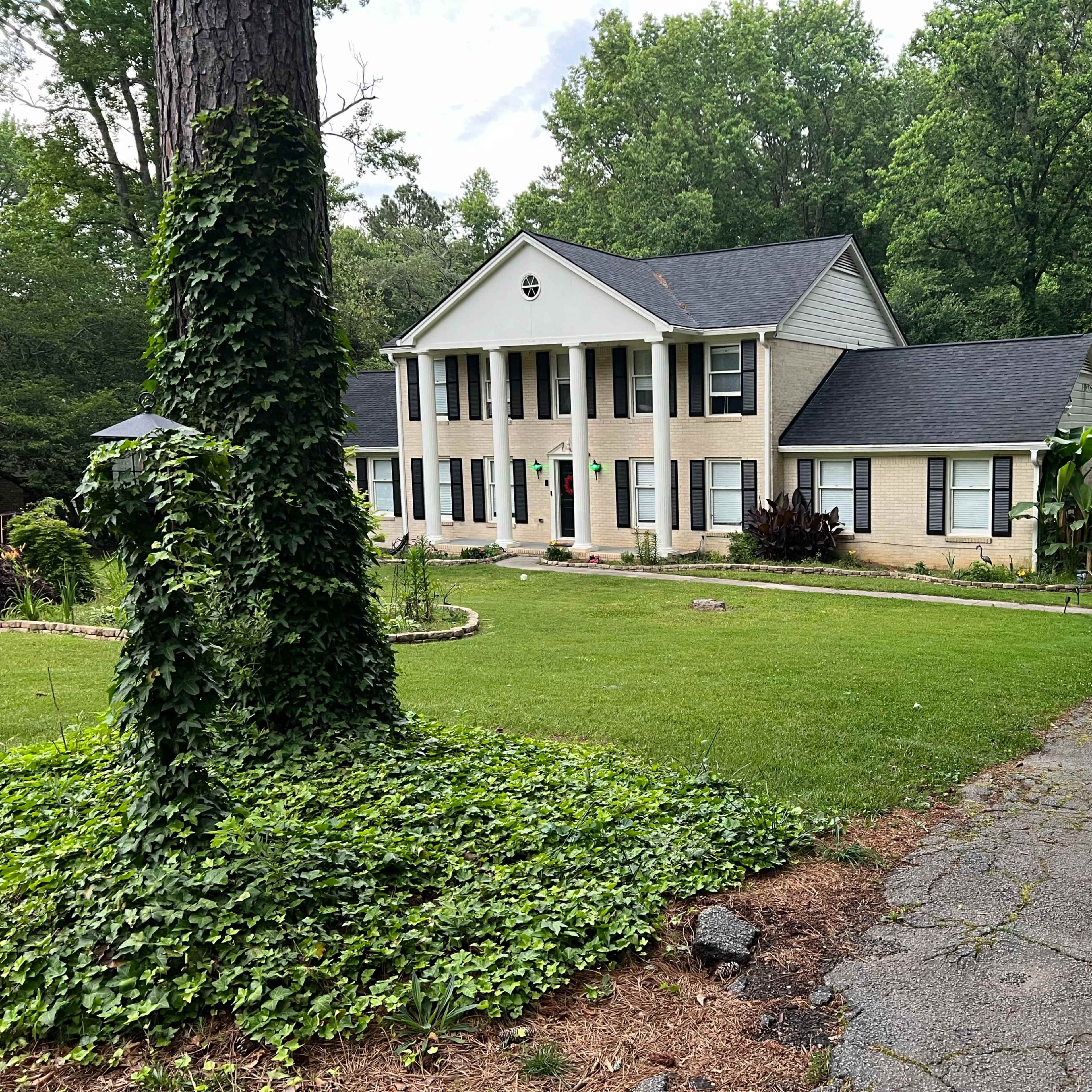 A two-story house with a light-colored facade and a large front lawn is surrounded by trees and a gravel driveway.