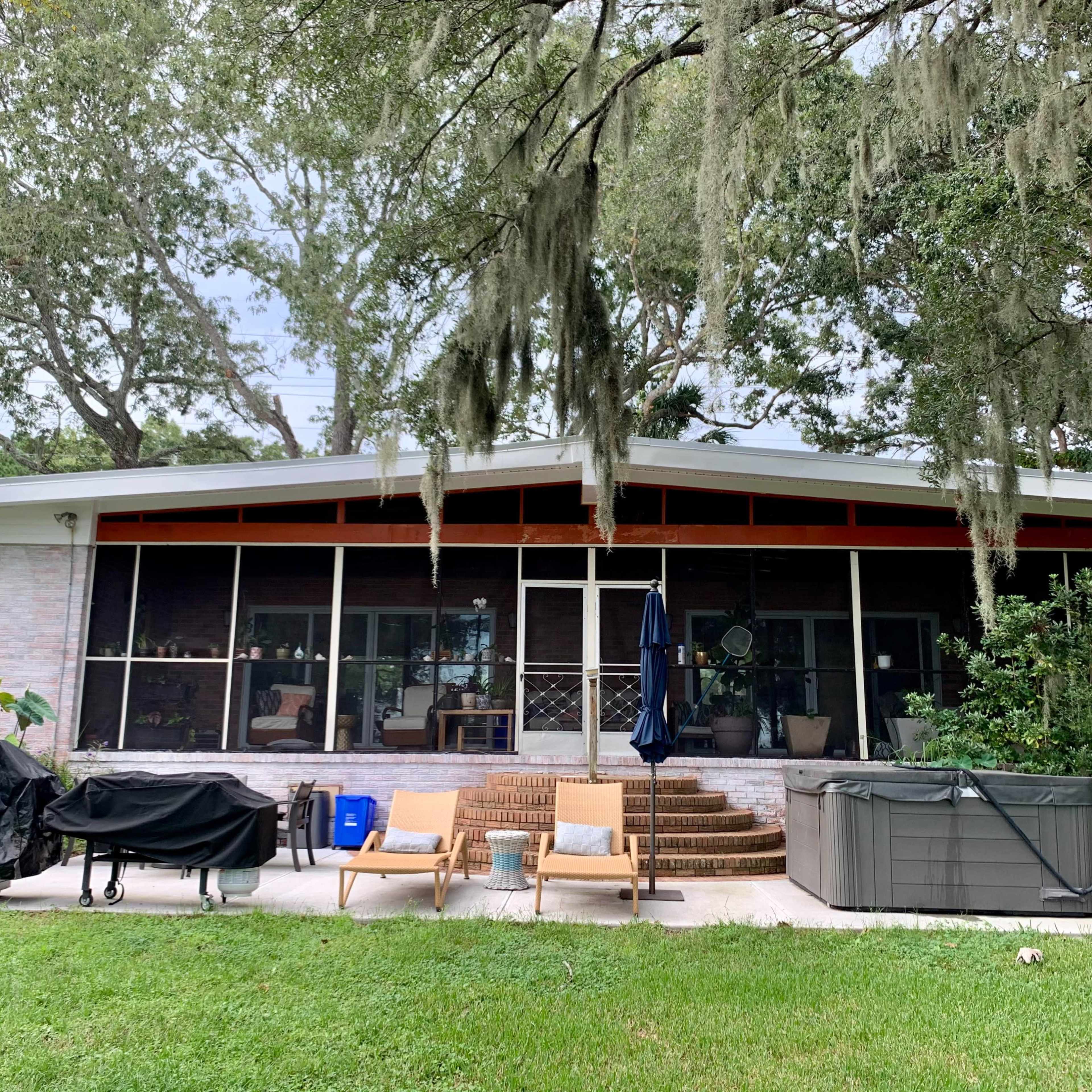A one-story house with a screened porch and outdoor seating, surrounded by trees and lawn space.