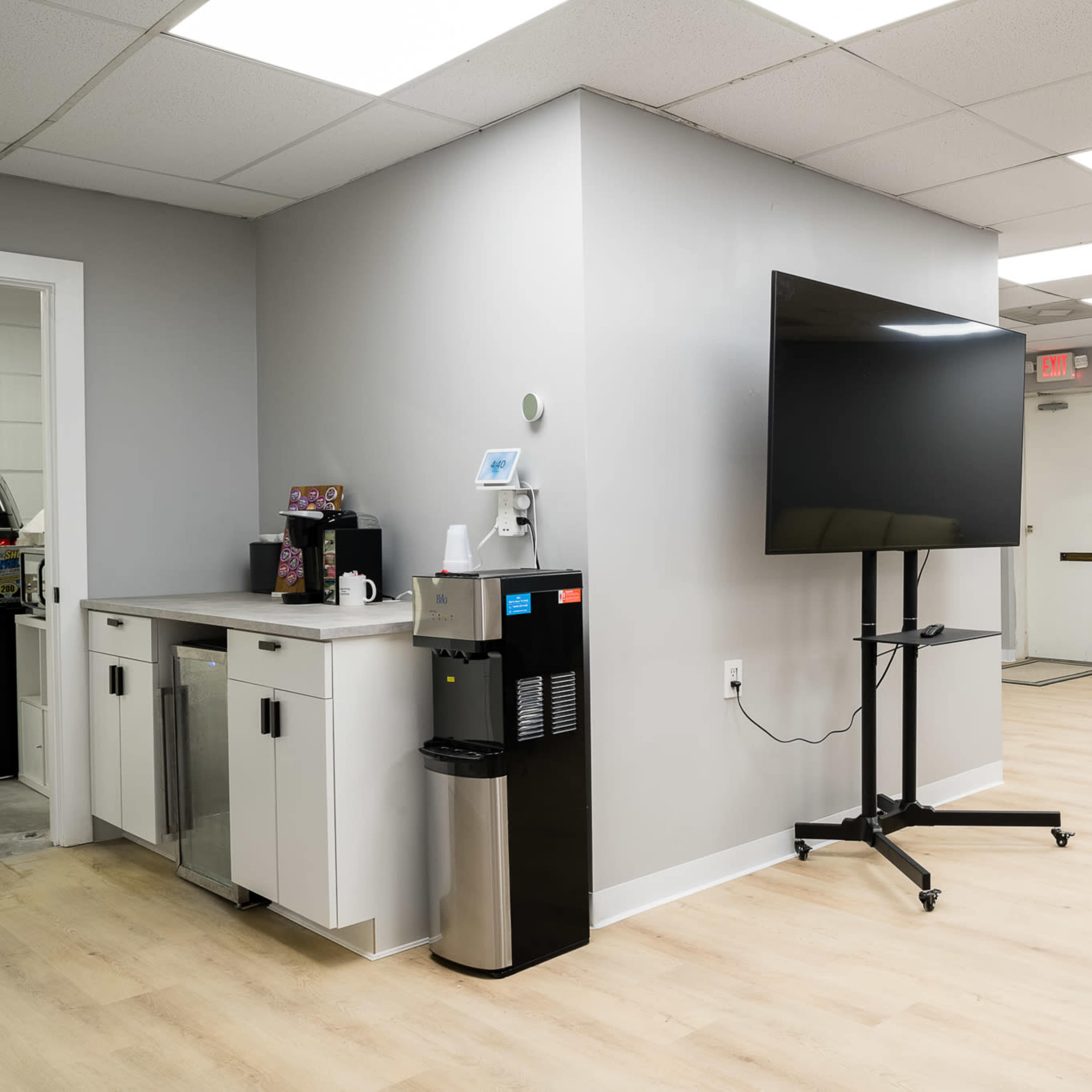 A modern office space with a kitchenette featuring a water cooler, a large television on a stand, and a seating area with a bench near a window.