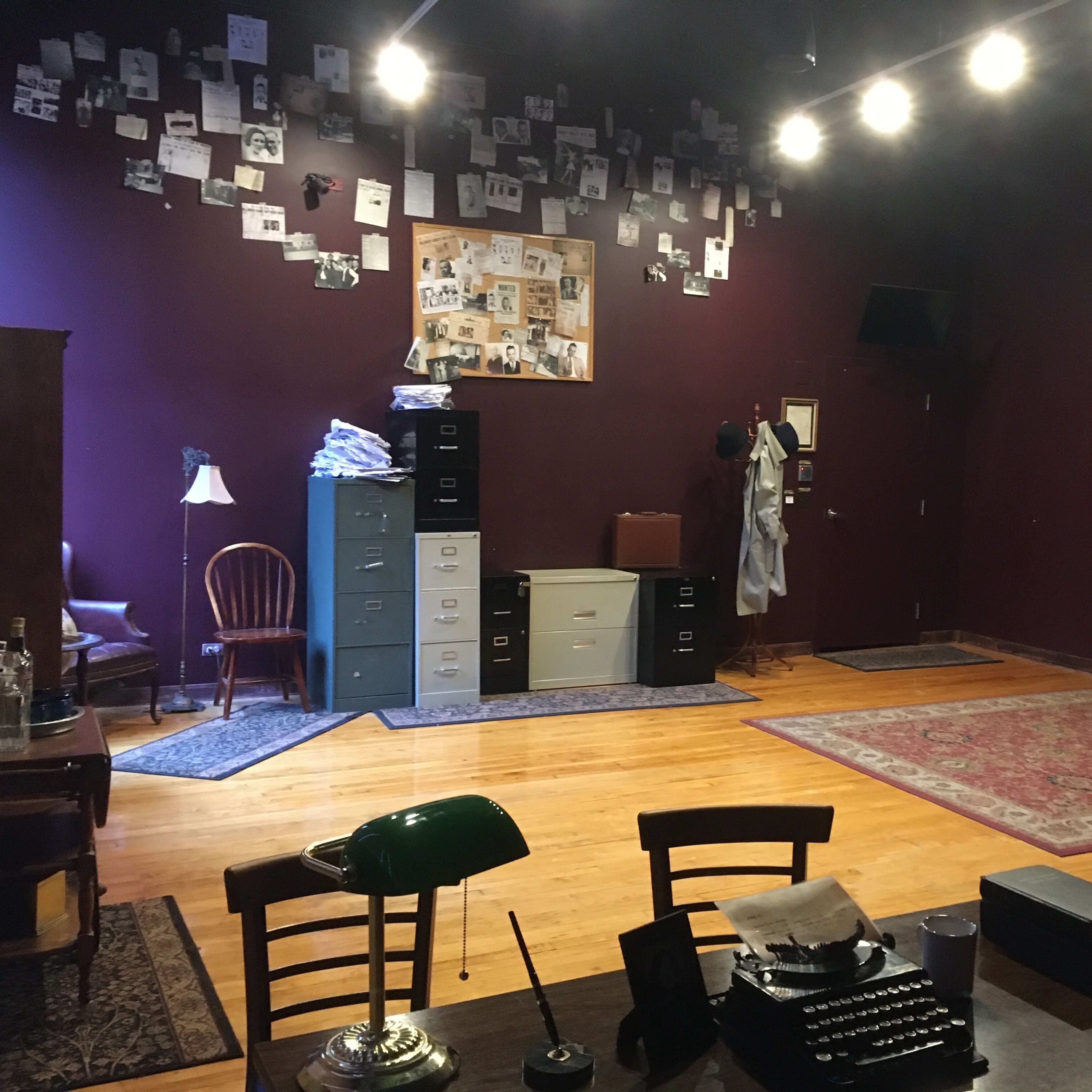 An office space with purple walls, wooden flooring, a desk with a green lamp and a typewriter, and filing cabinets against the back wall adorned with black-and-white photos.