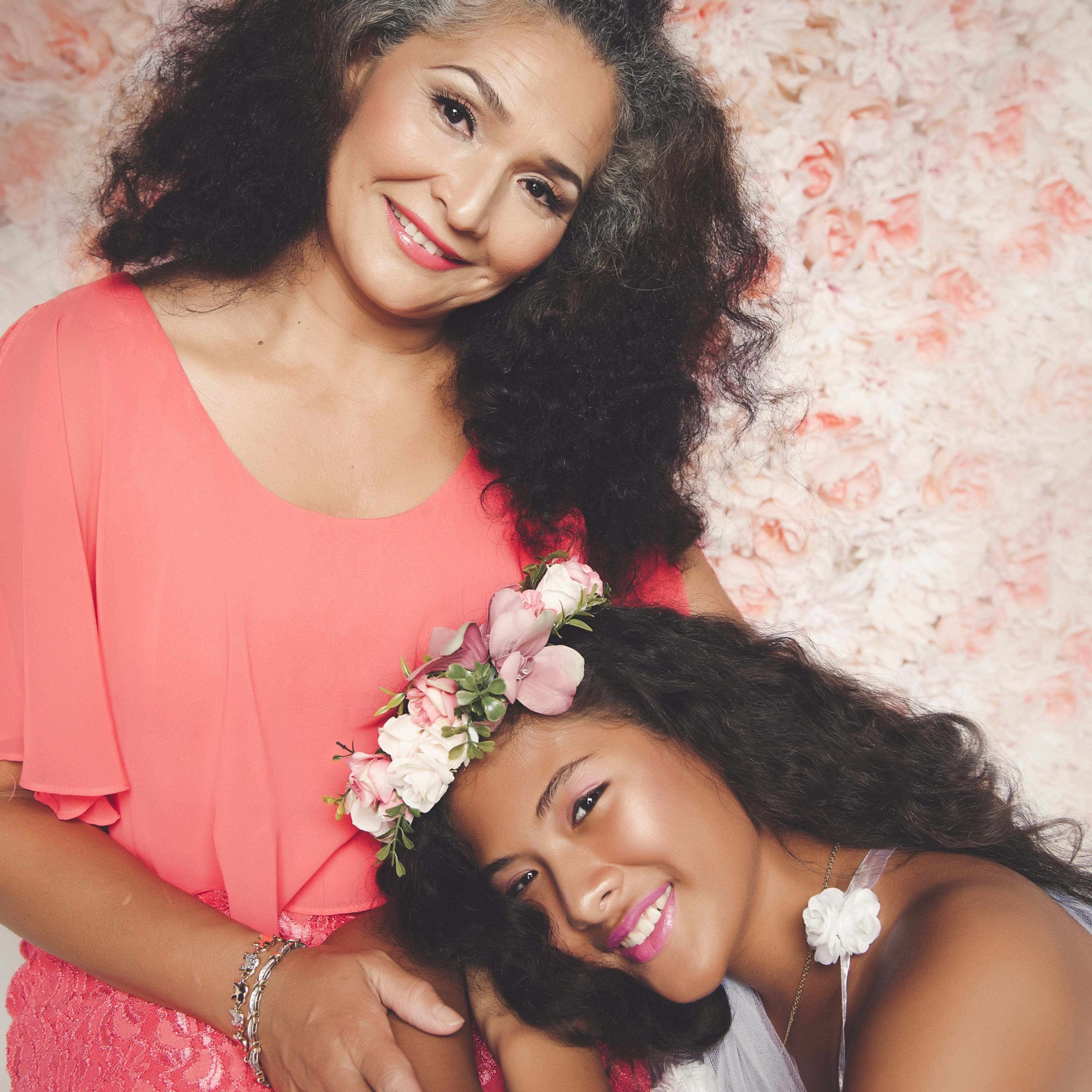 A woman in a pink blouse poses with a girl wearing a flower crown, both smiling against a floral backdrop.