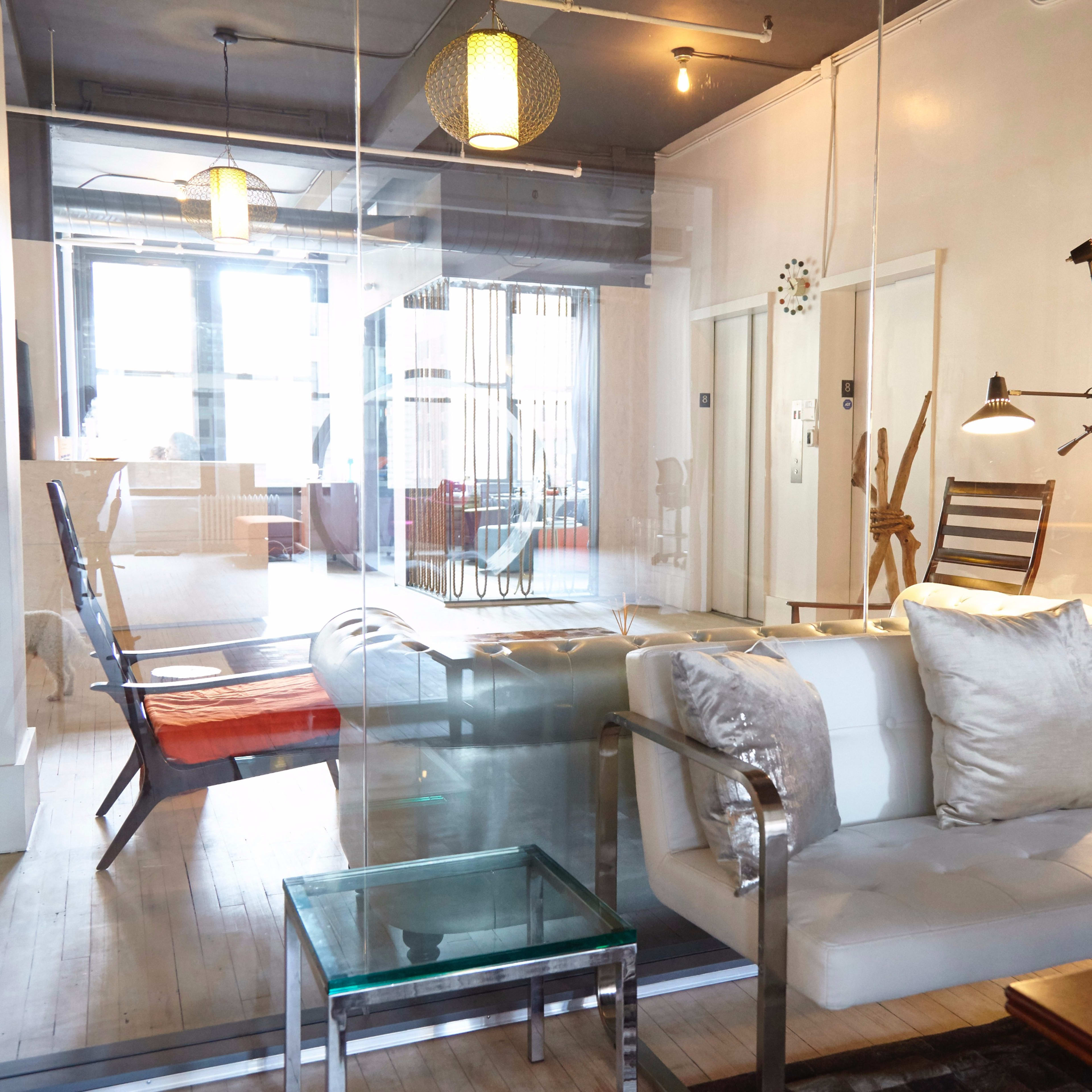 A modern office space features a glass partition separating a lounge area with a white couch and a table from a well-lit room with wooden furniture.