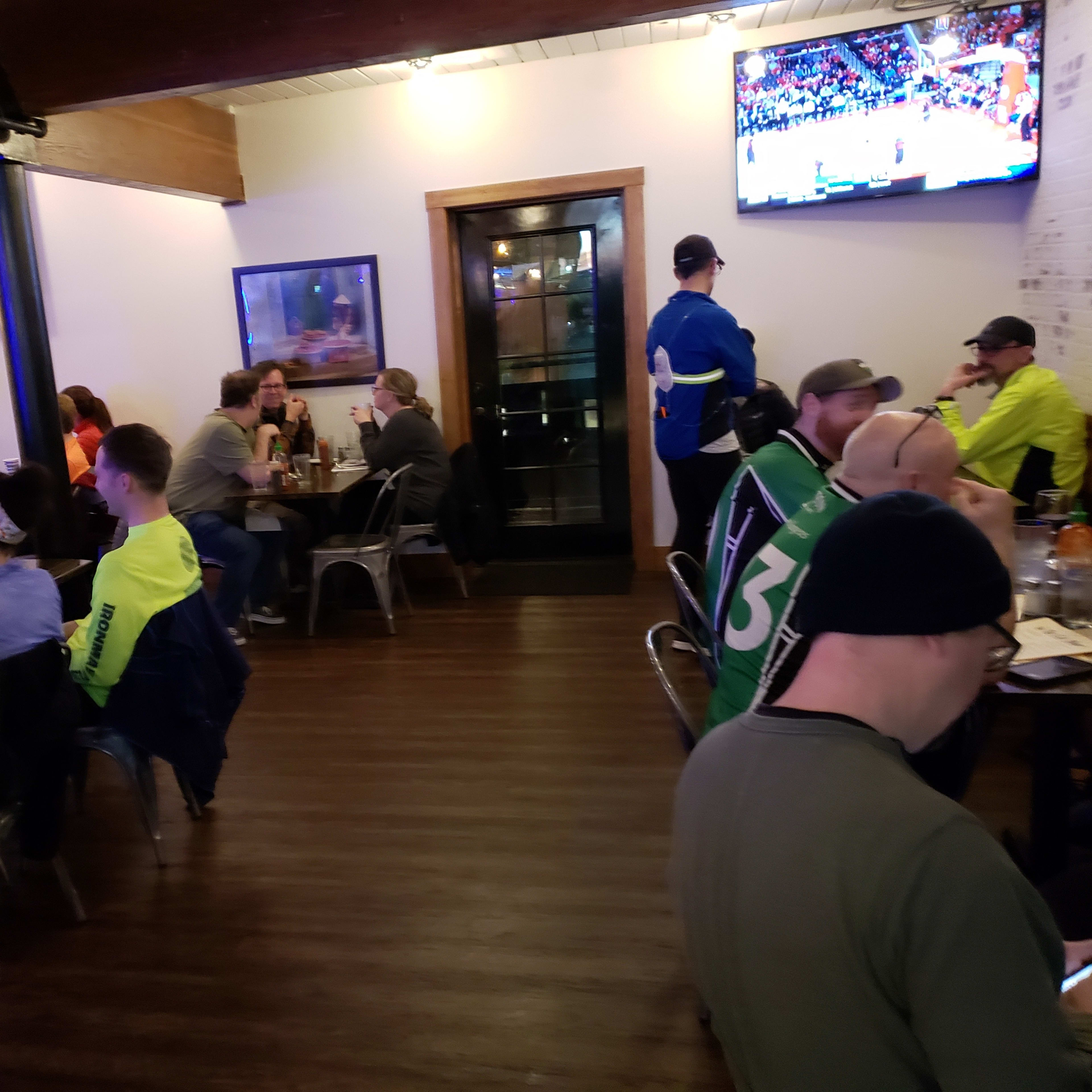 A restaurant interior with patrons sitting at tables, a staff member taking an order, and multiple televisions displaying a sports event.