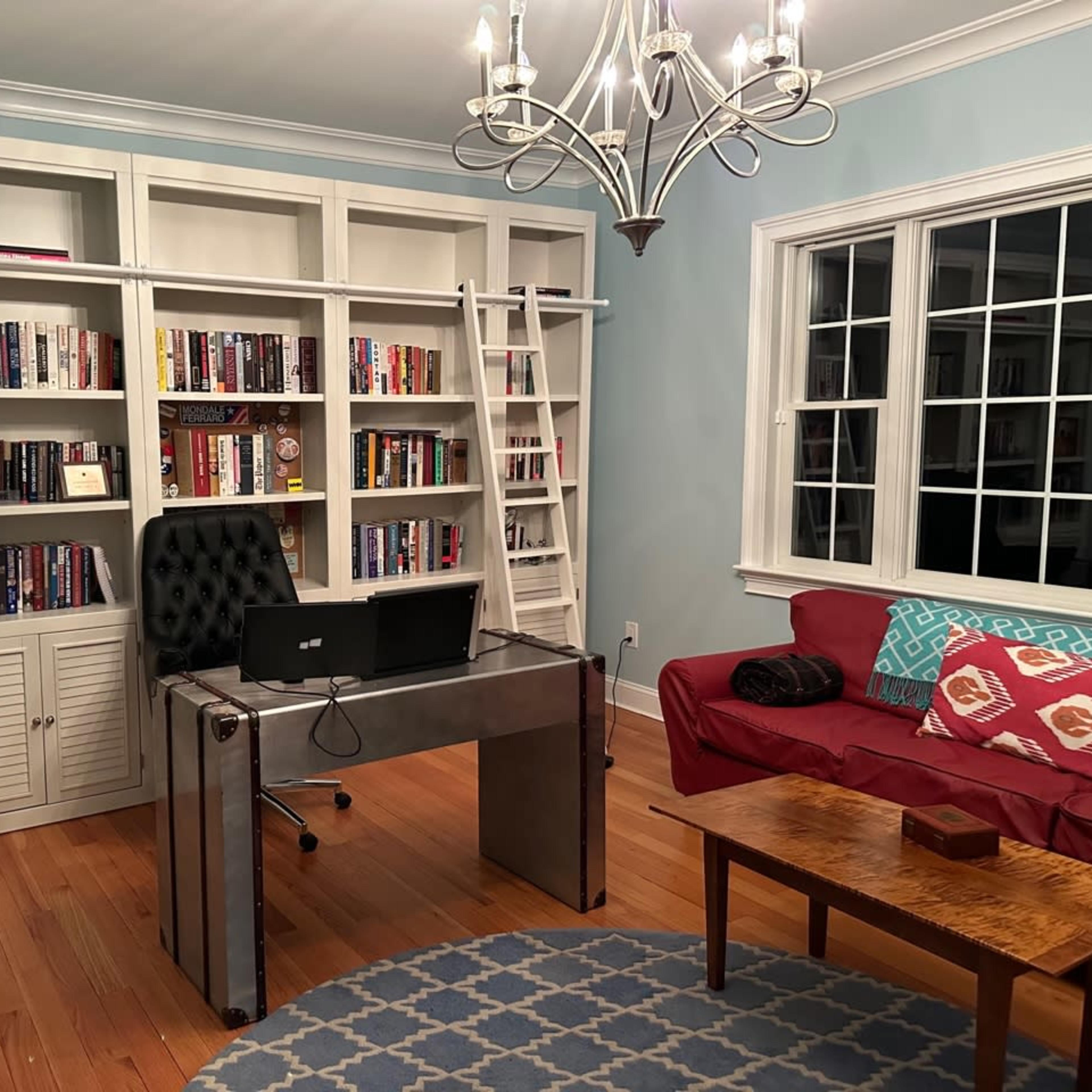 A home office with a metal desk, a computer monitor, a red couch, and a bookshelf filled with books.