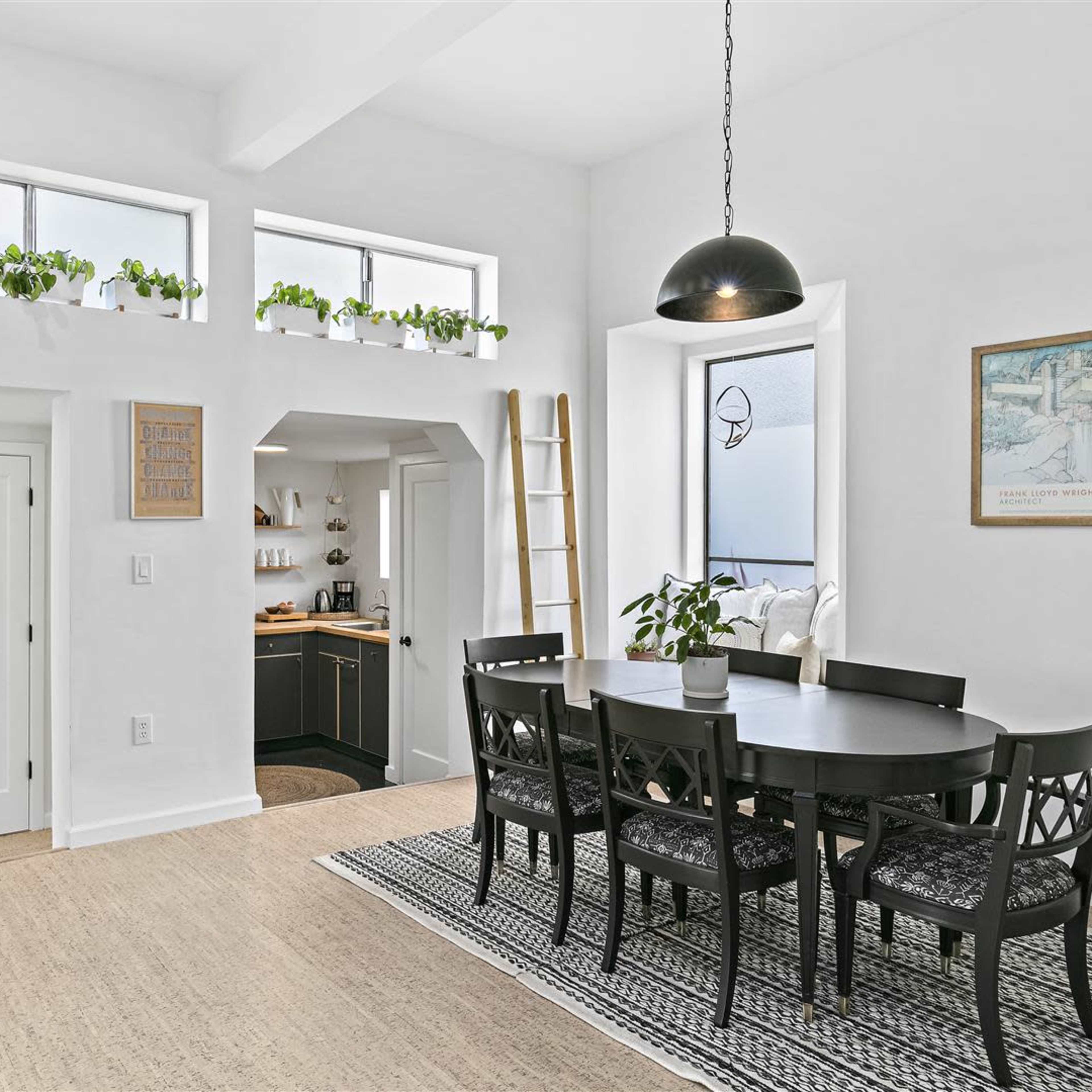 A bright dining area featuring a round table surrounded by black chairs, with a kitchen visible in the background and greenery in window planters.