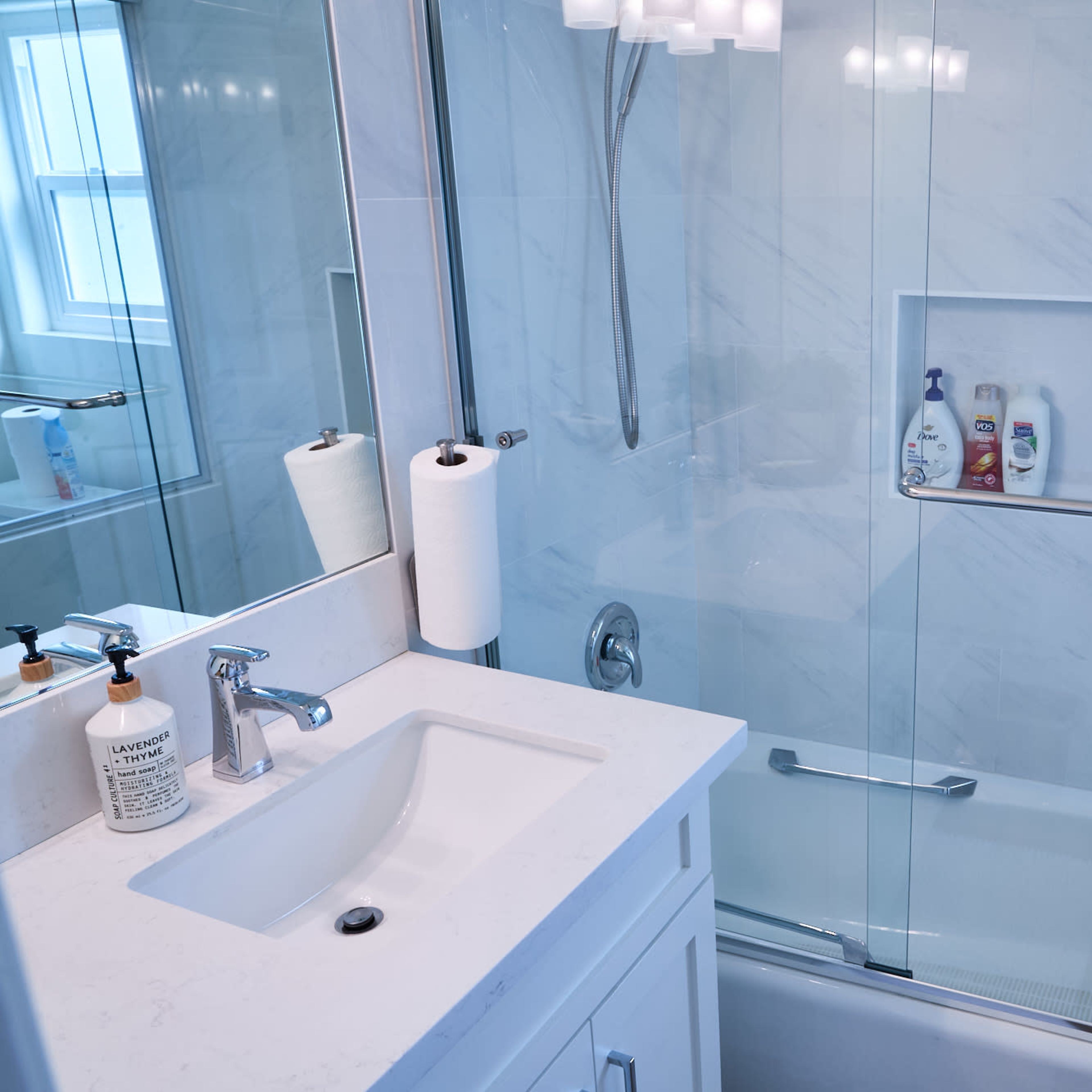 A modern bathroom with a glass shower enclosure, a white countertop with a sink, and toiletries displayed on the shelves.