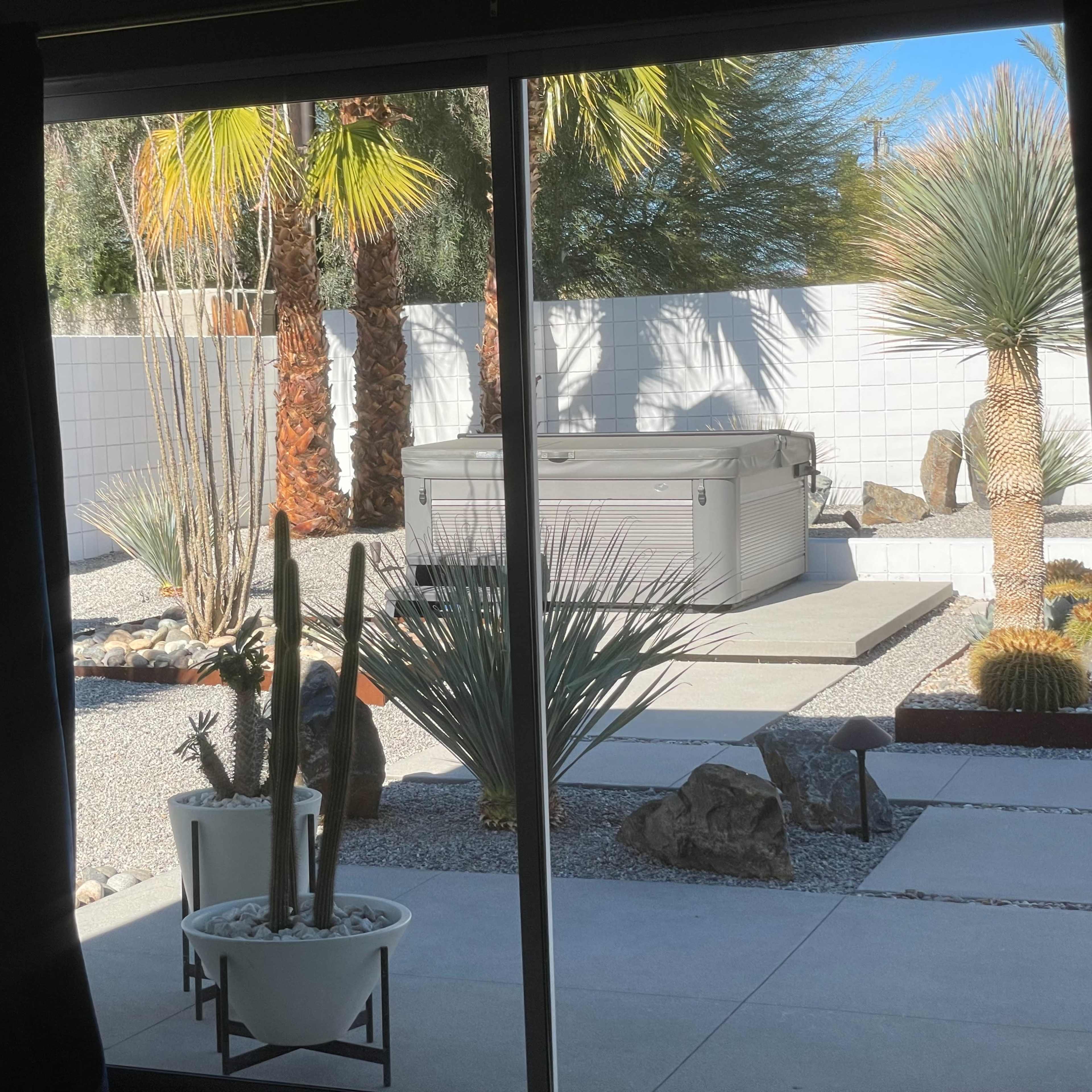 A view through a sliding glass door of a landscaped backyard featuring a hot tub, various desert plants, and gravel pathways.
