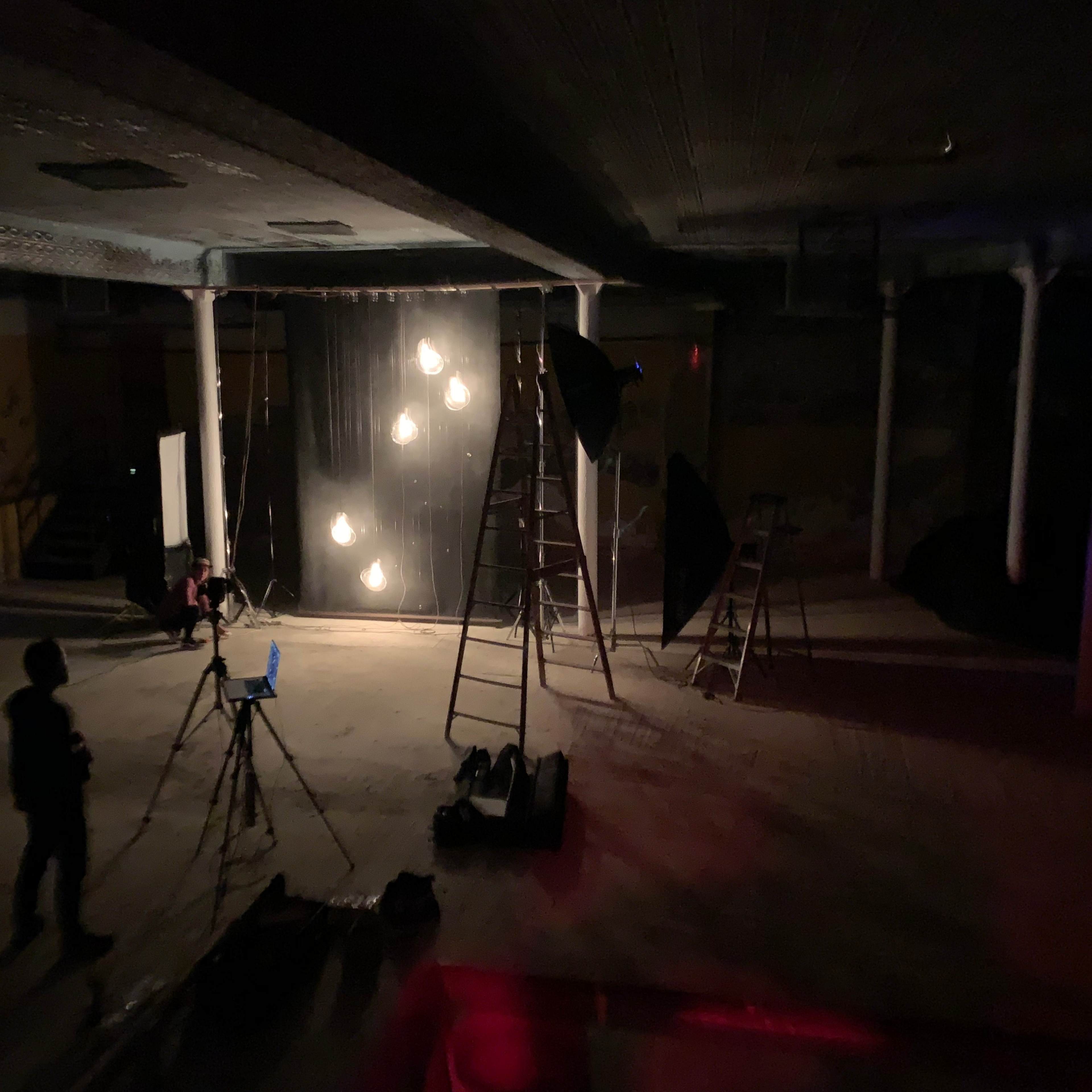 A dimly lit space features ladders, lighting equipment, and a camera set up in front of a backdrop with hanging lights.