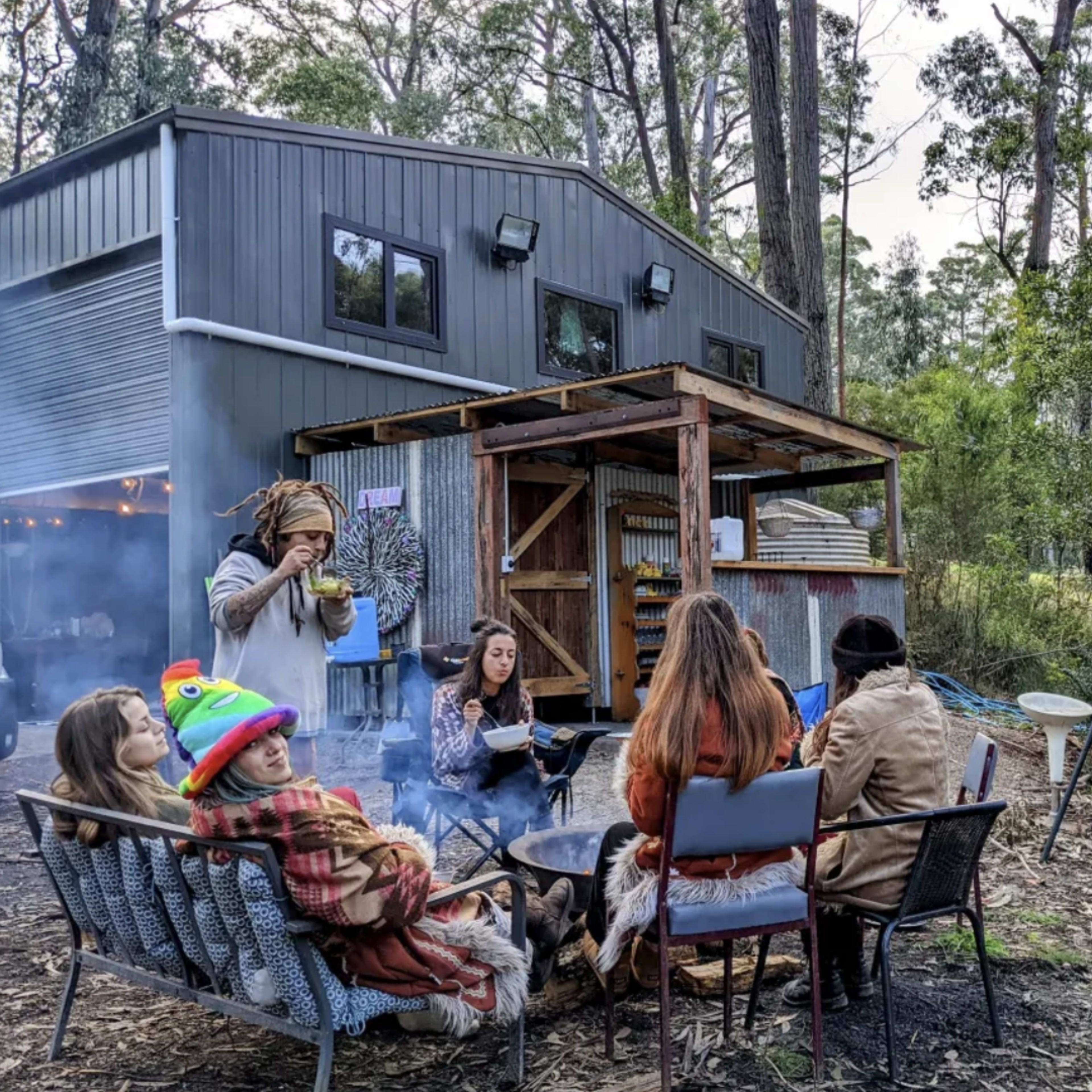 A group of people sits around a fire pit in a wooded area, with a modern cabin visible in the background.