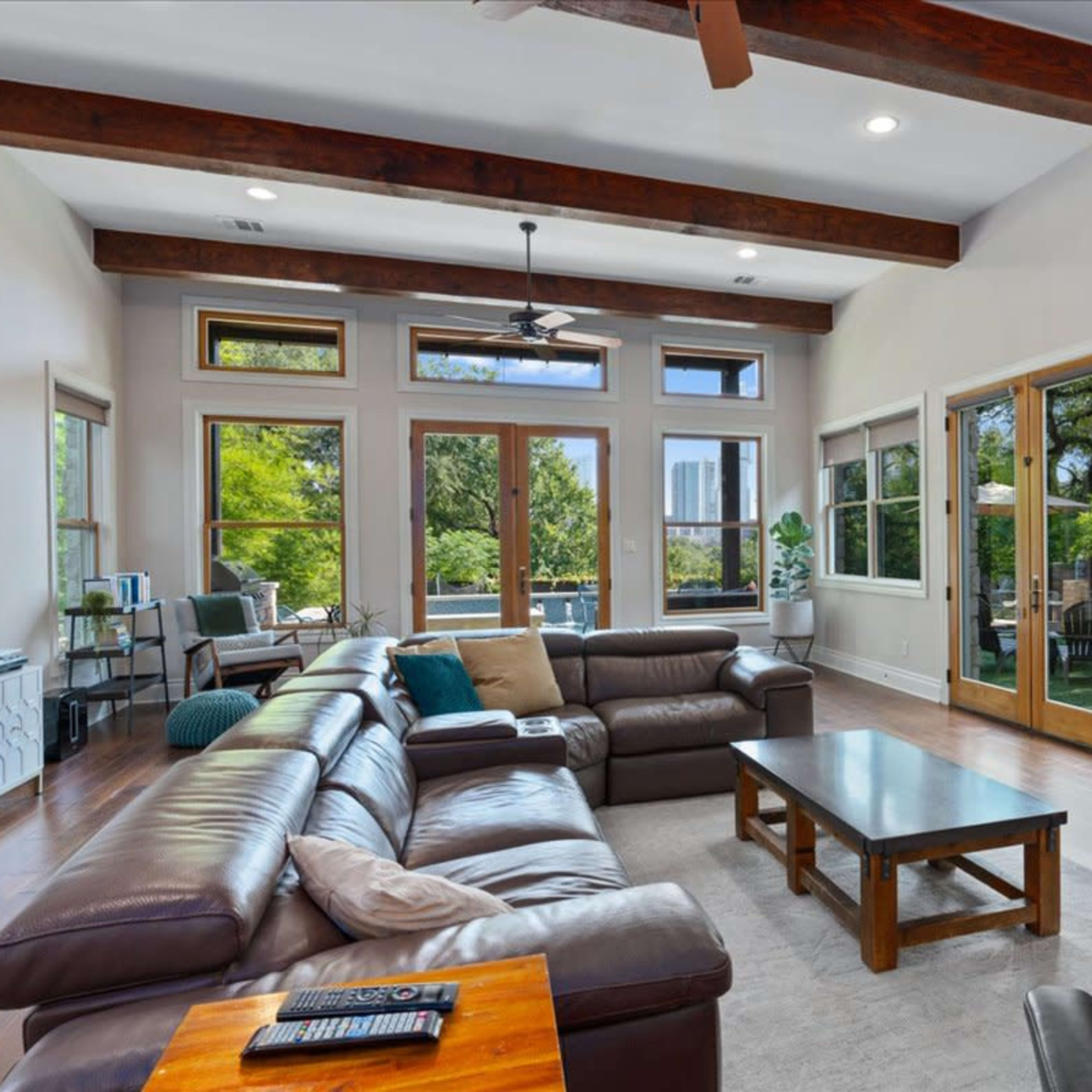 A spacious living room features a leather sectional sofa, a coffee table, large windows with views, and a mounted television on a stone wall.