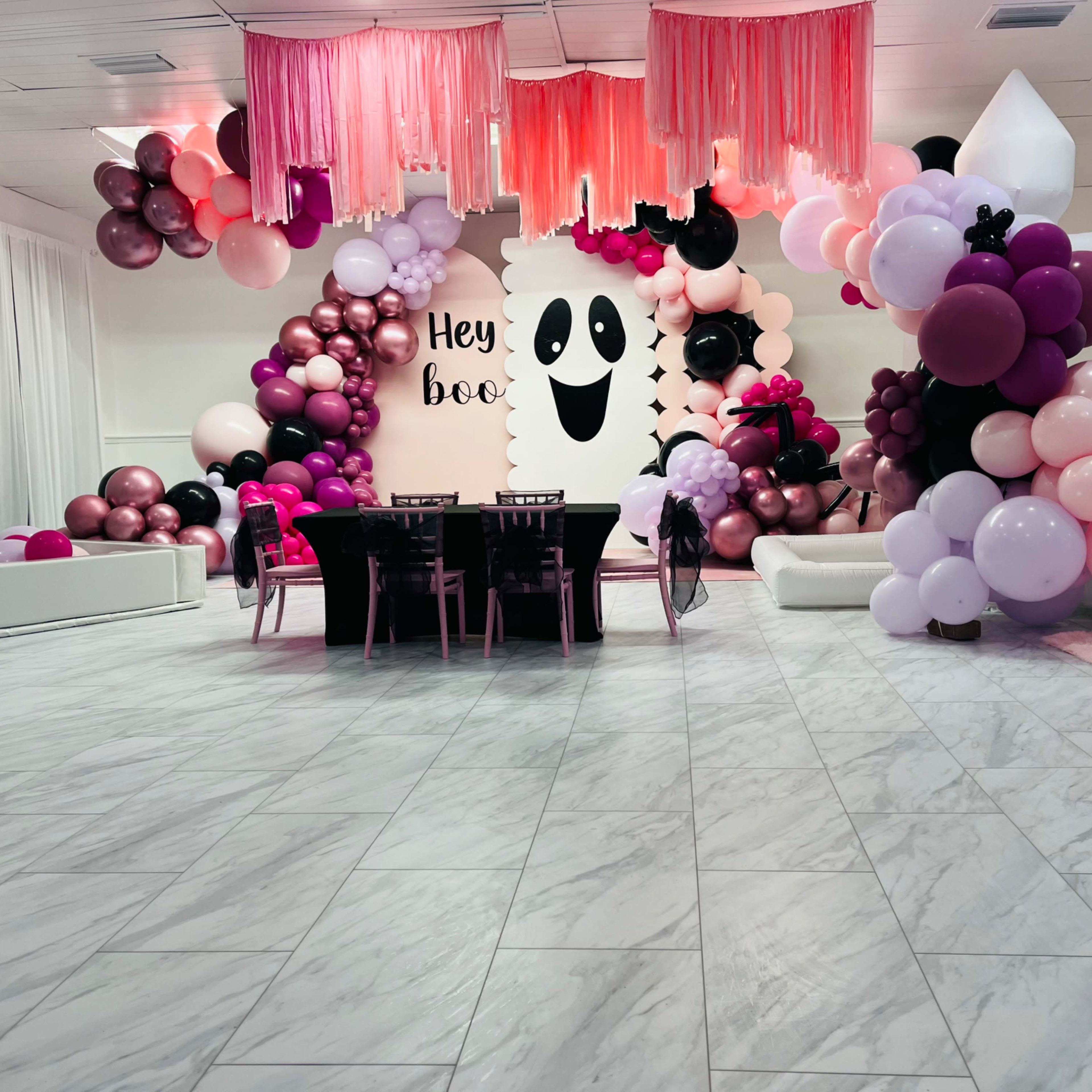 A decorated event space featuring a table set for guests, surrounded by an assortment of balloons in various colors and a wall with a ghost-themed design that reads "Hey boo."