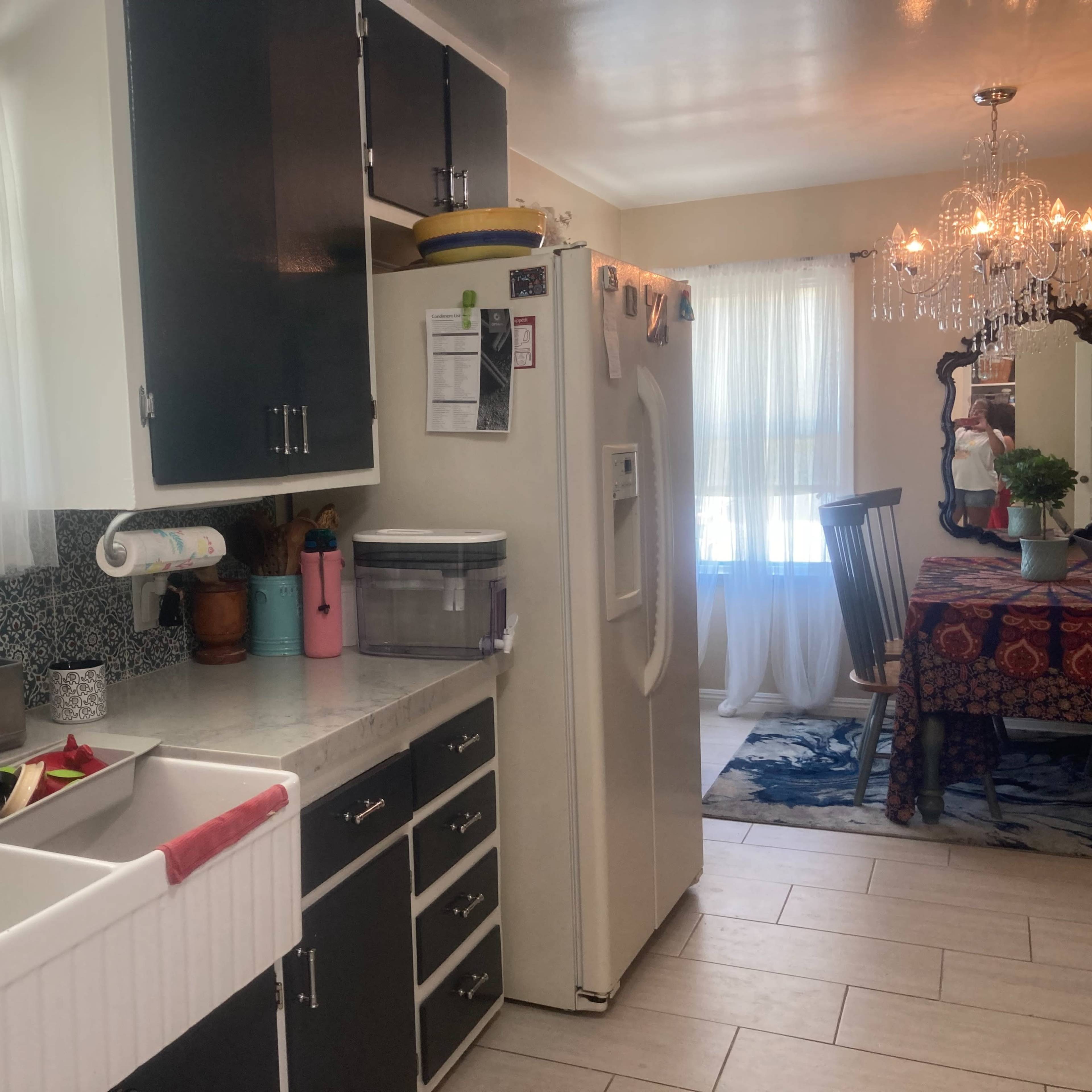 A kitchen with a farmhouse sink, black cabinets, a white refrigerator, and a dining area visible through the doorway.