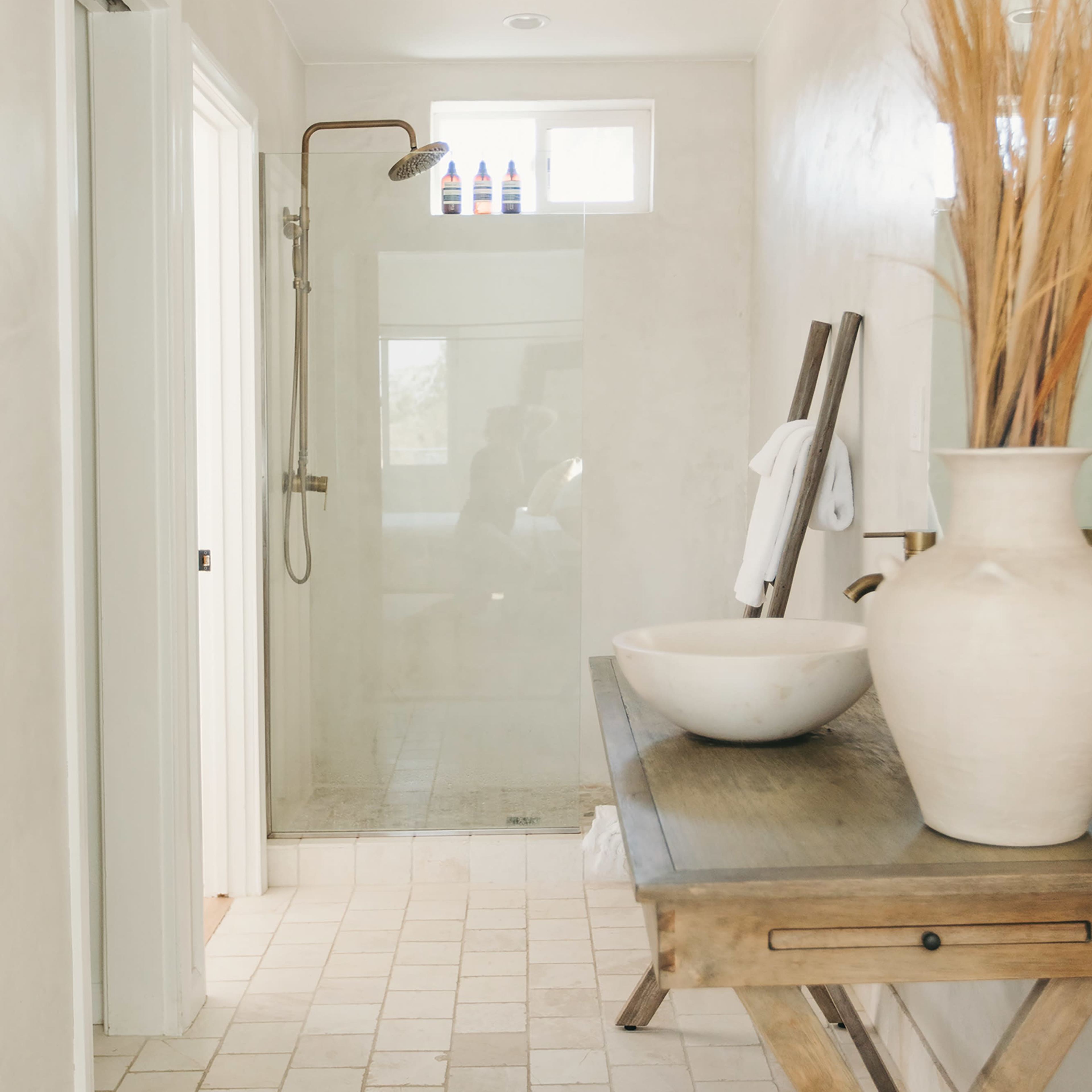 A modern bathroom features a glass shower, a wooden table with a vase, and a neutral color palette.