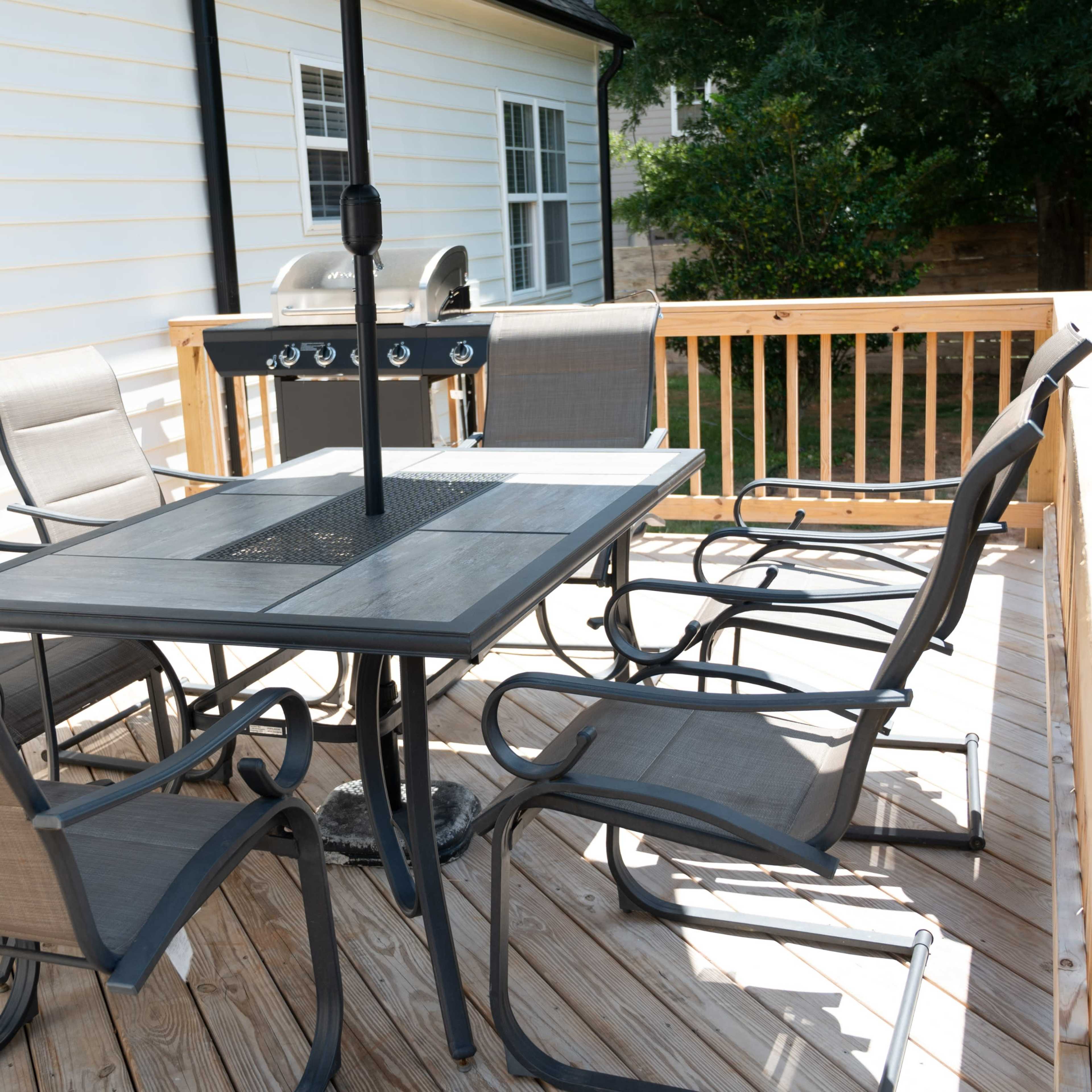 A six-seat outdoor dining table with an umbrella and a grill is positioned on a wooden deck.