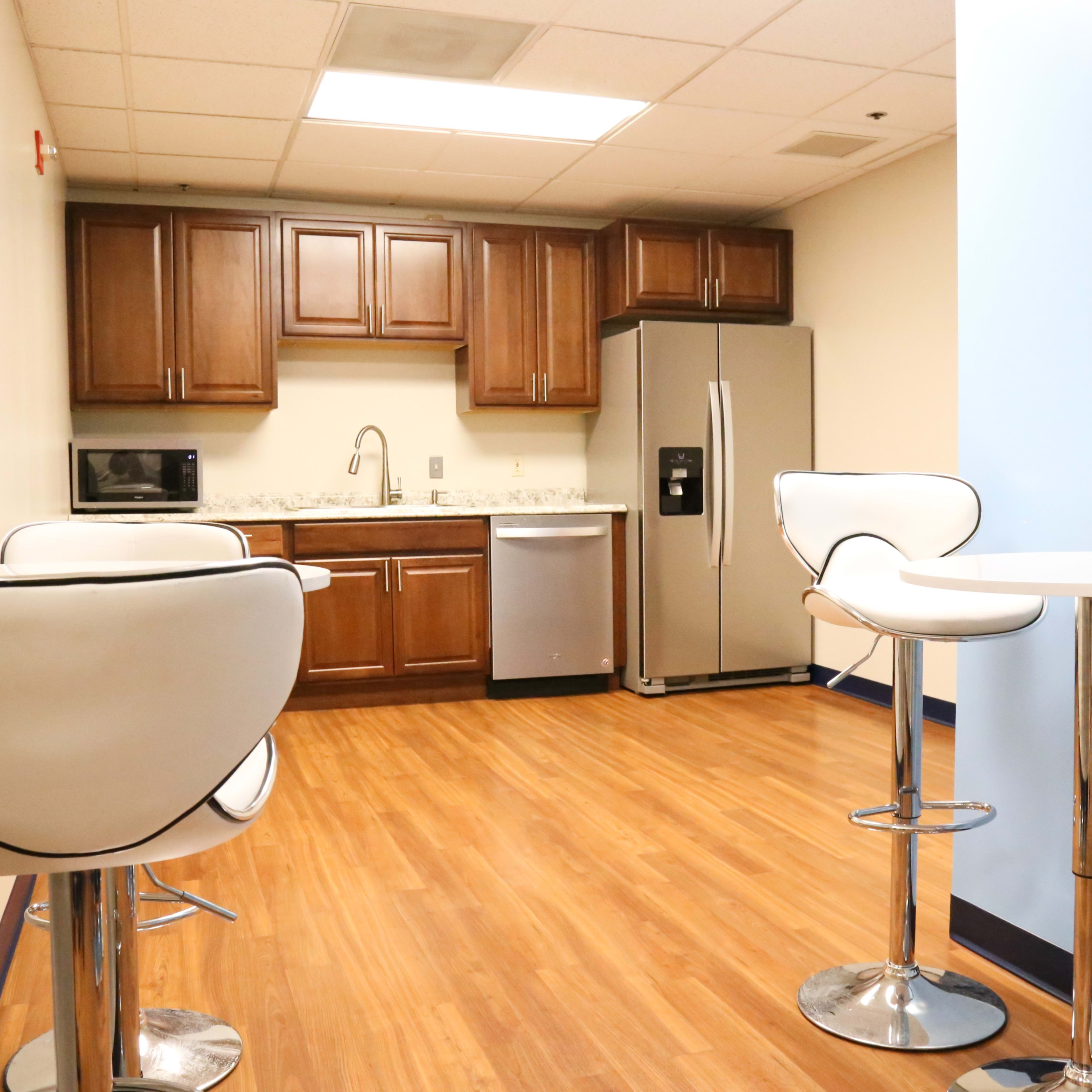 A modern kitchenette with brown cabinets, stainless steel appliances, and white bar stools around circular tables.