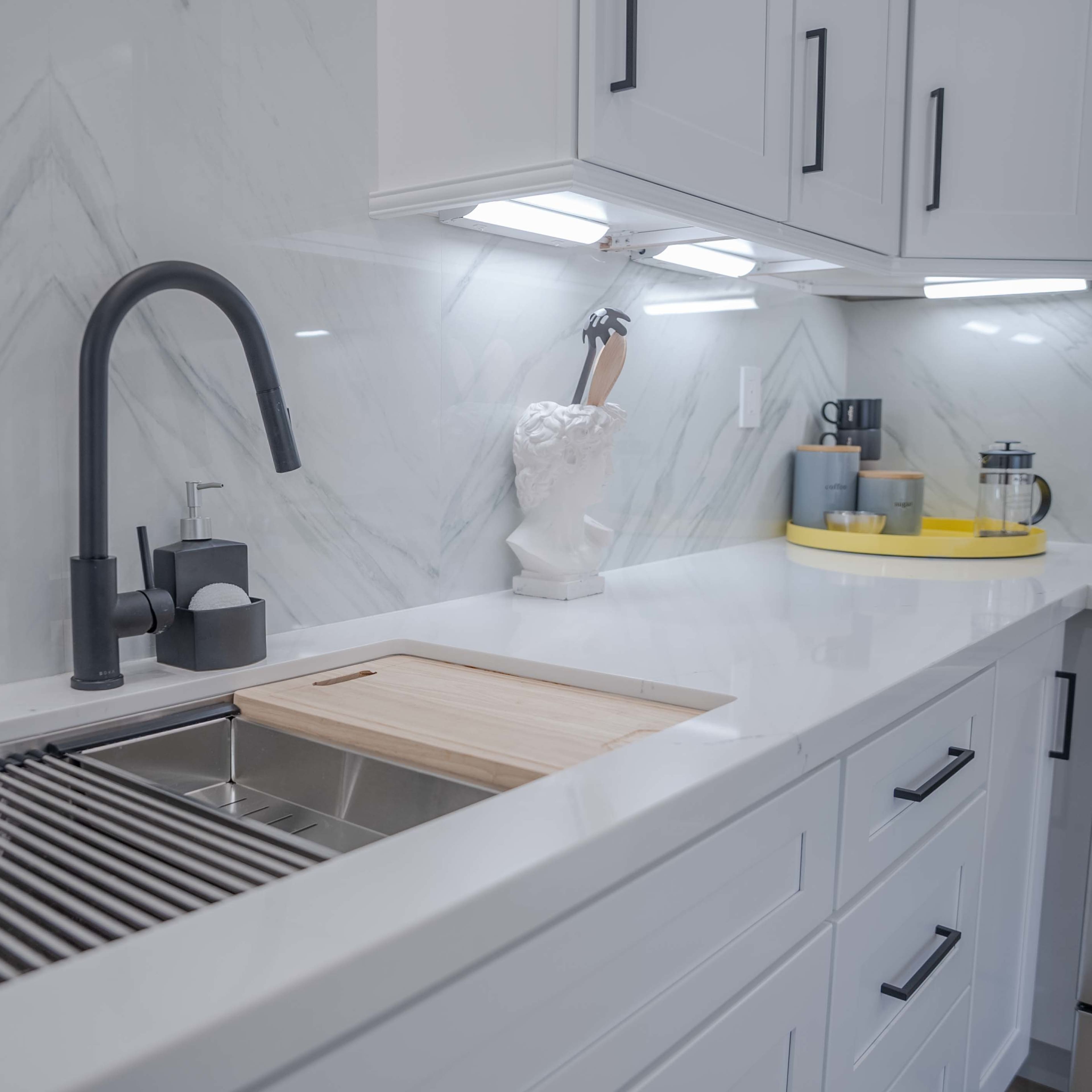 A modern kitchen sink with a wooden cutting board, a decorative bust, and a counter with various kitchen utensils and a coffee maker.