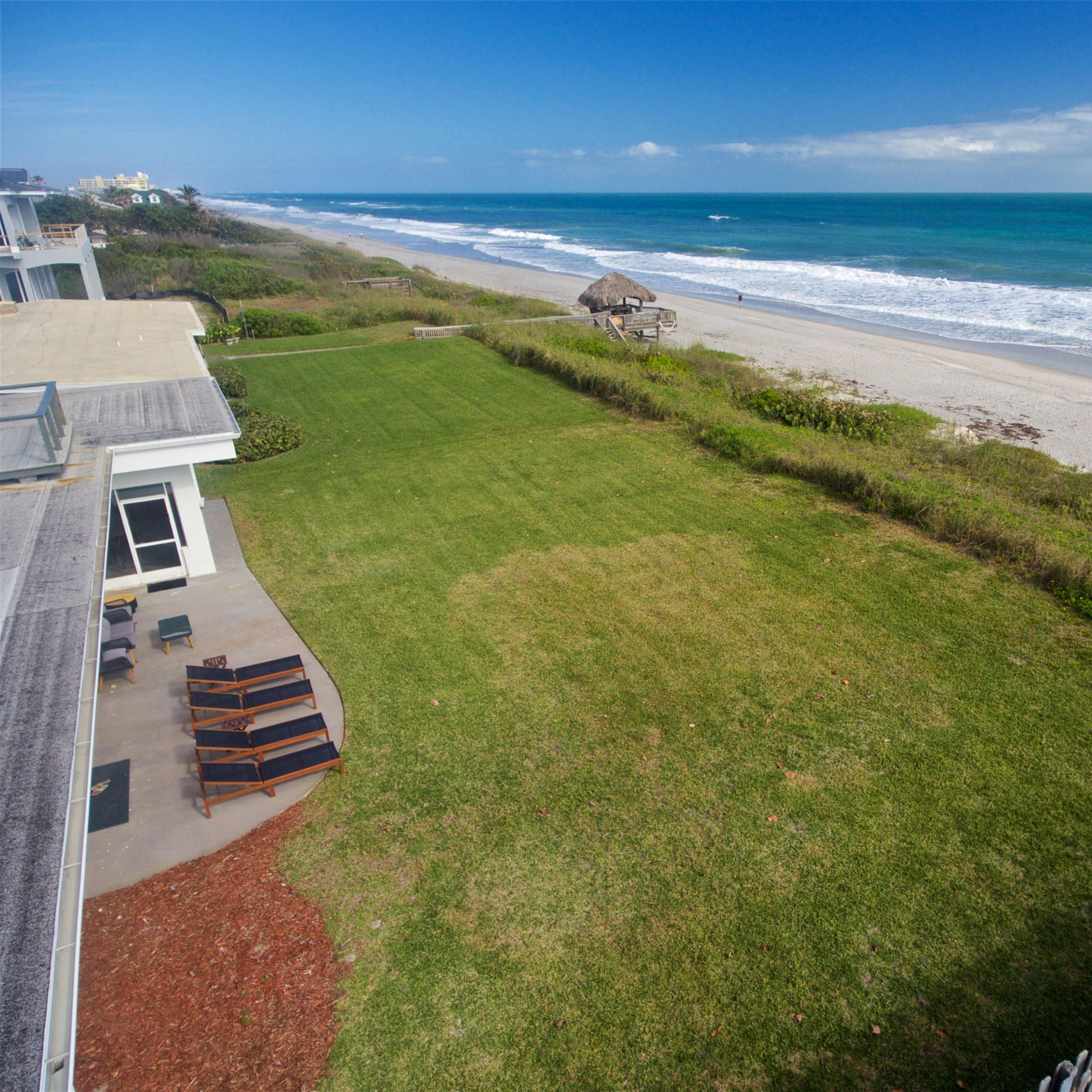 A coastal landscape with a beachfront lawn, a sandy shore, and a few people walking along the water's edge.