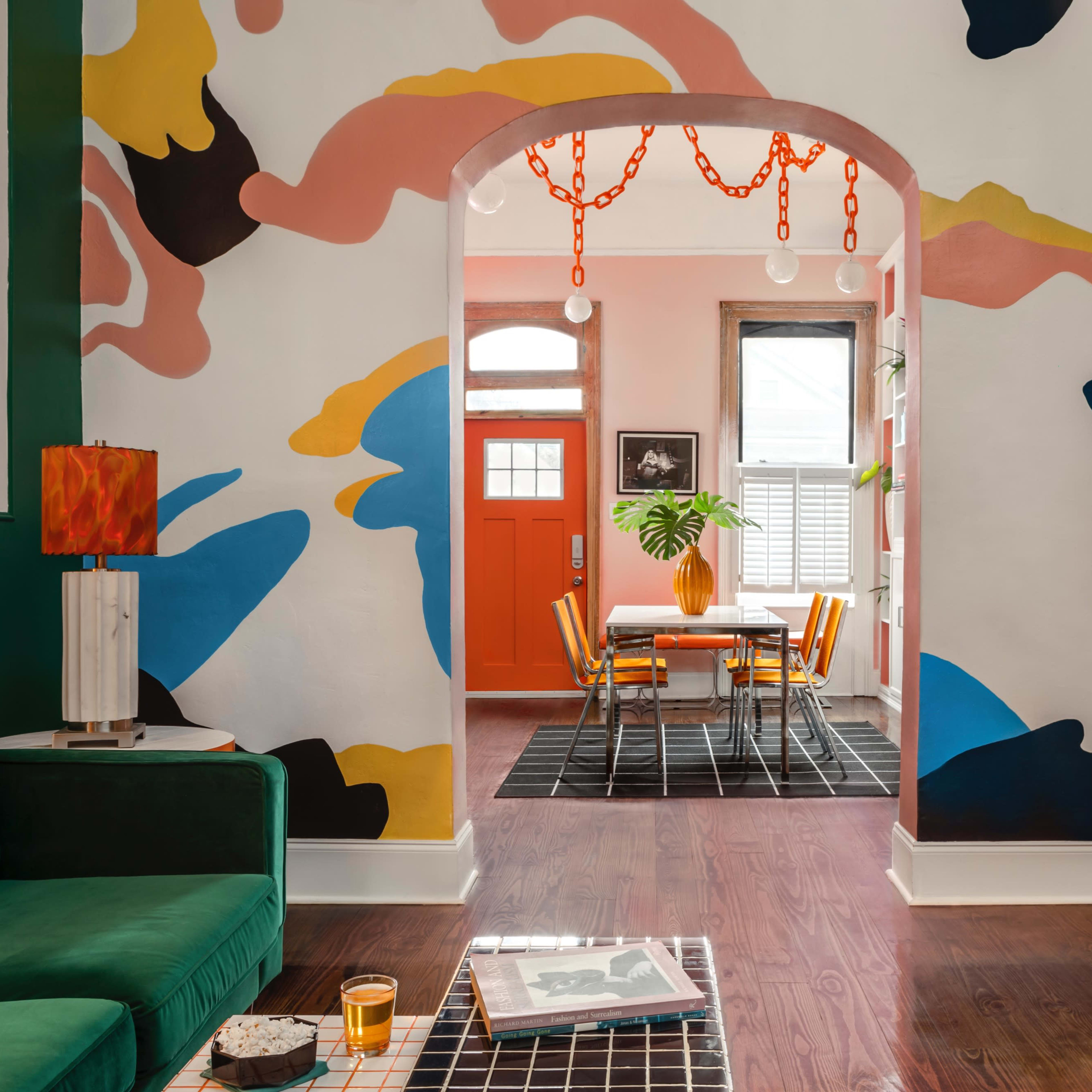 A colorful living room features a green couch, a bold mural on the wall, and an orange door leading to a dining area with a table and chairs.