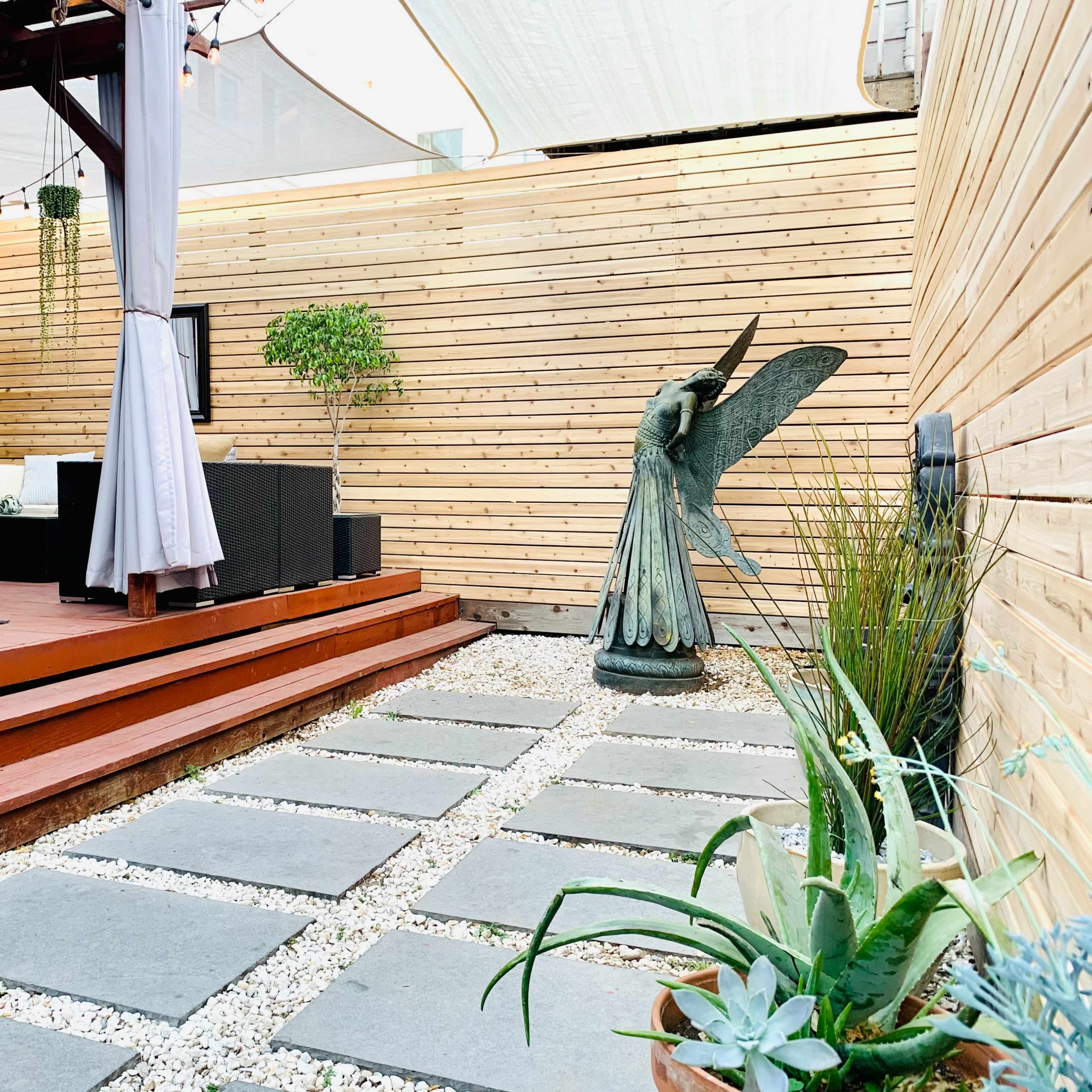 A modern outdoor patio area with wooden walls, a covered seating arrangement, and a stone pathway leading to a decorative angel statue.