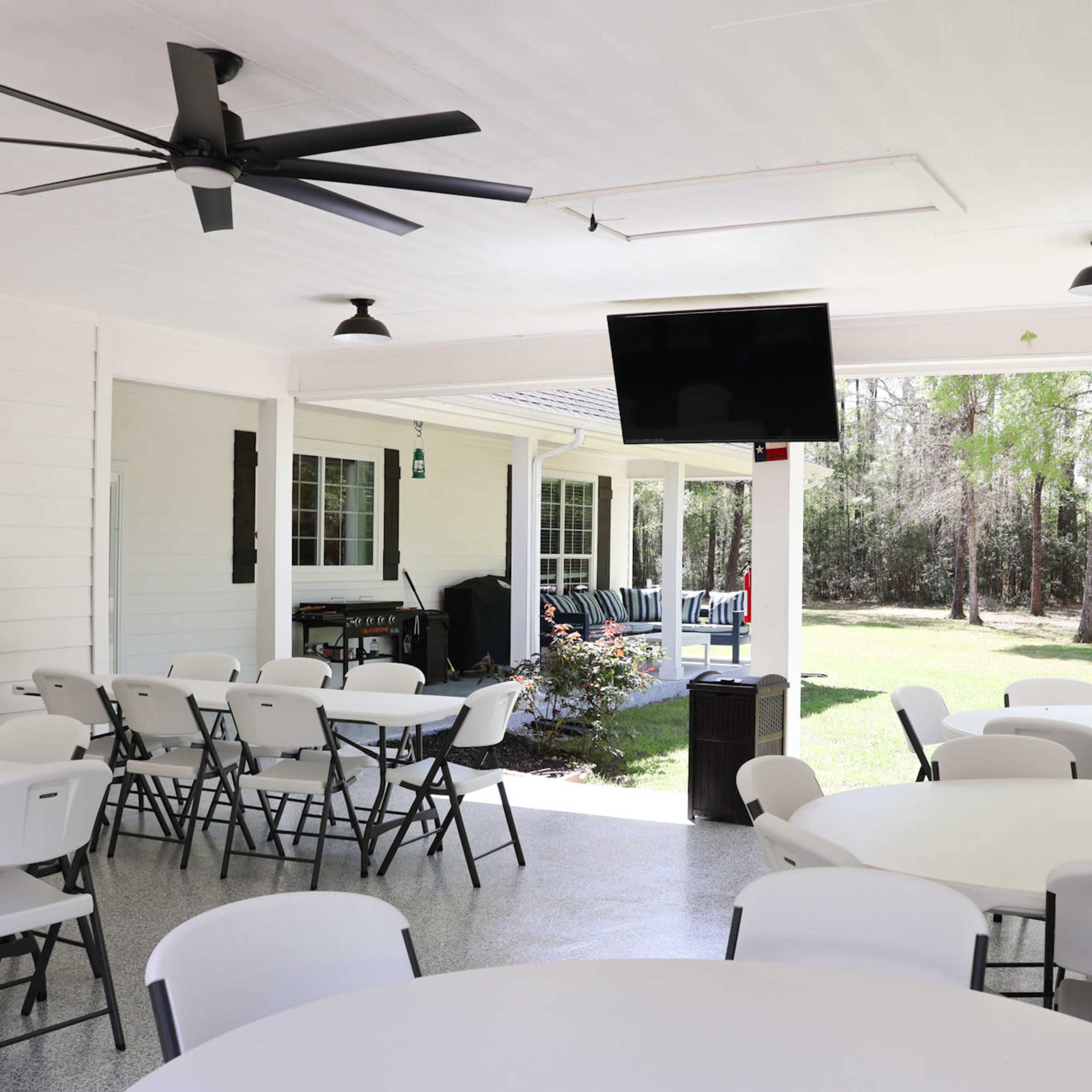 A spacious outdoor covered area with several circular tables and chairs, a ceiling fan, and a television mounted on the wall, overlooking a grassy yard with trees.