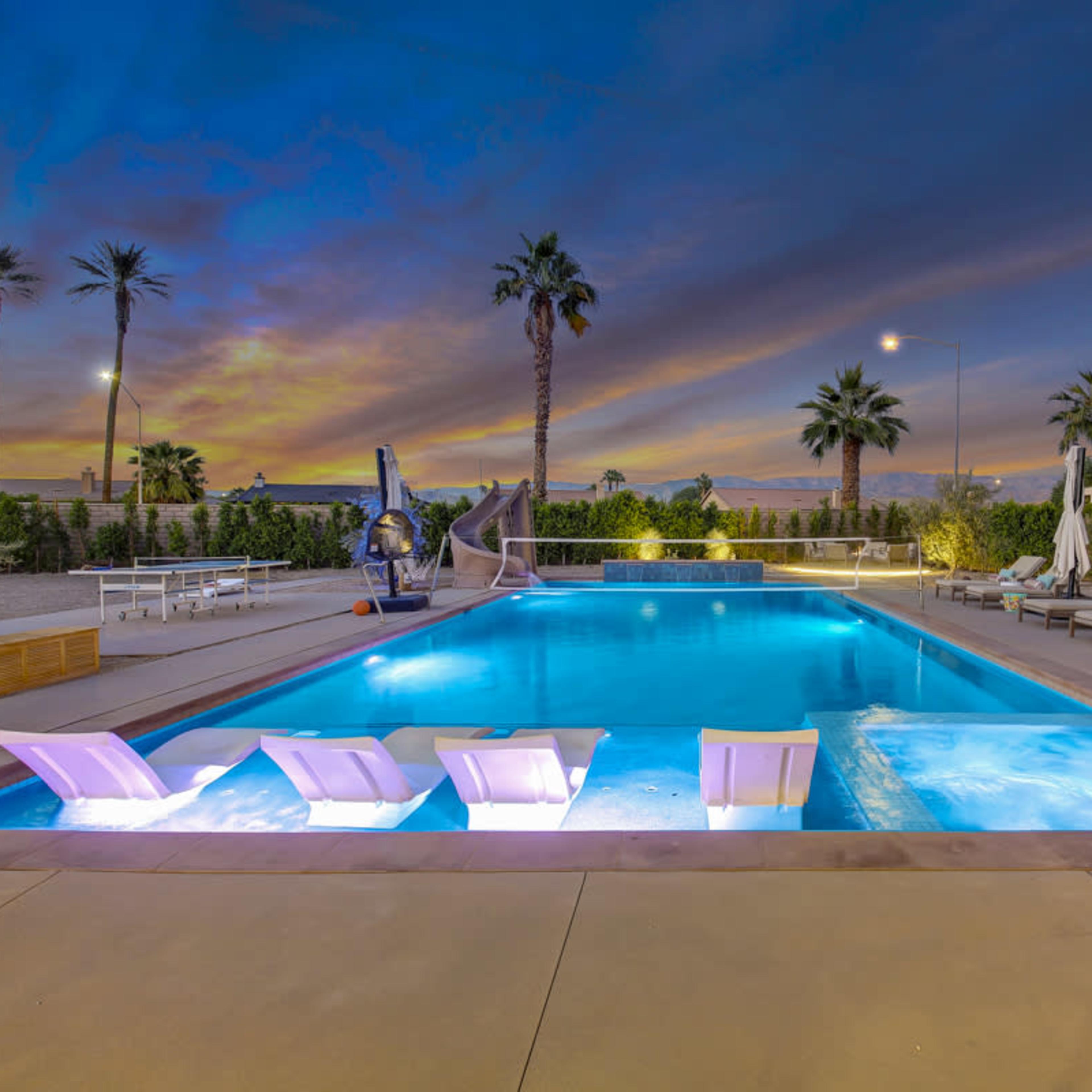 A luxurious outdoor pool area features lounge chairs, a hot tub, and palm trees under a dusk sky.